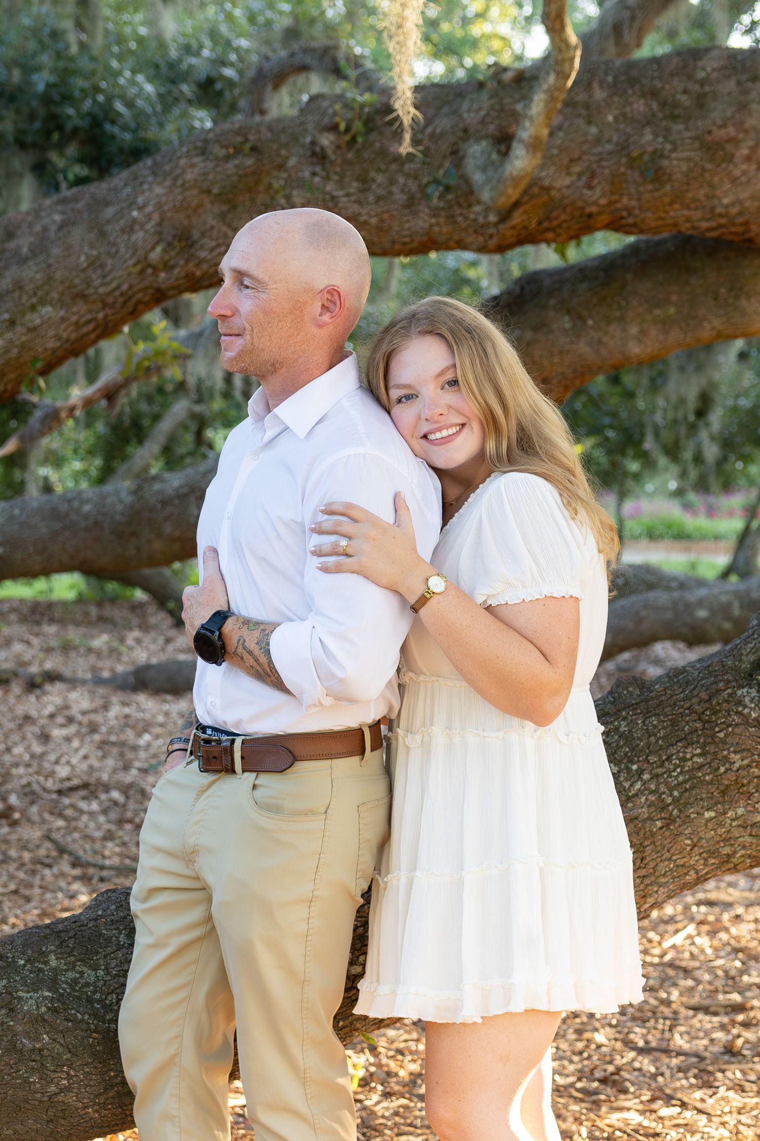 A smiling woman in a white dress embraces a bald man in a white shirt outdoors near a large oak tree.