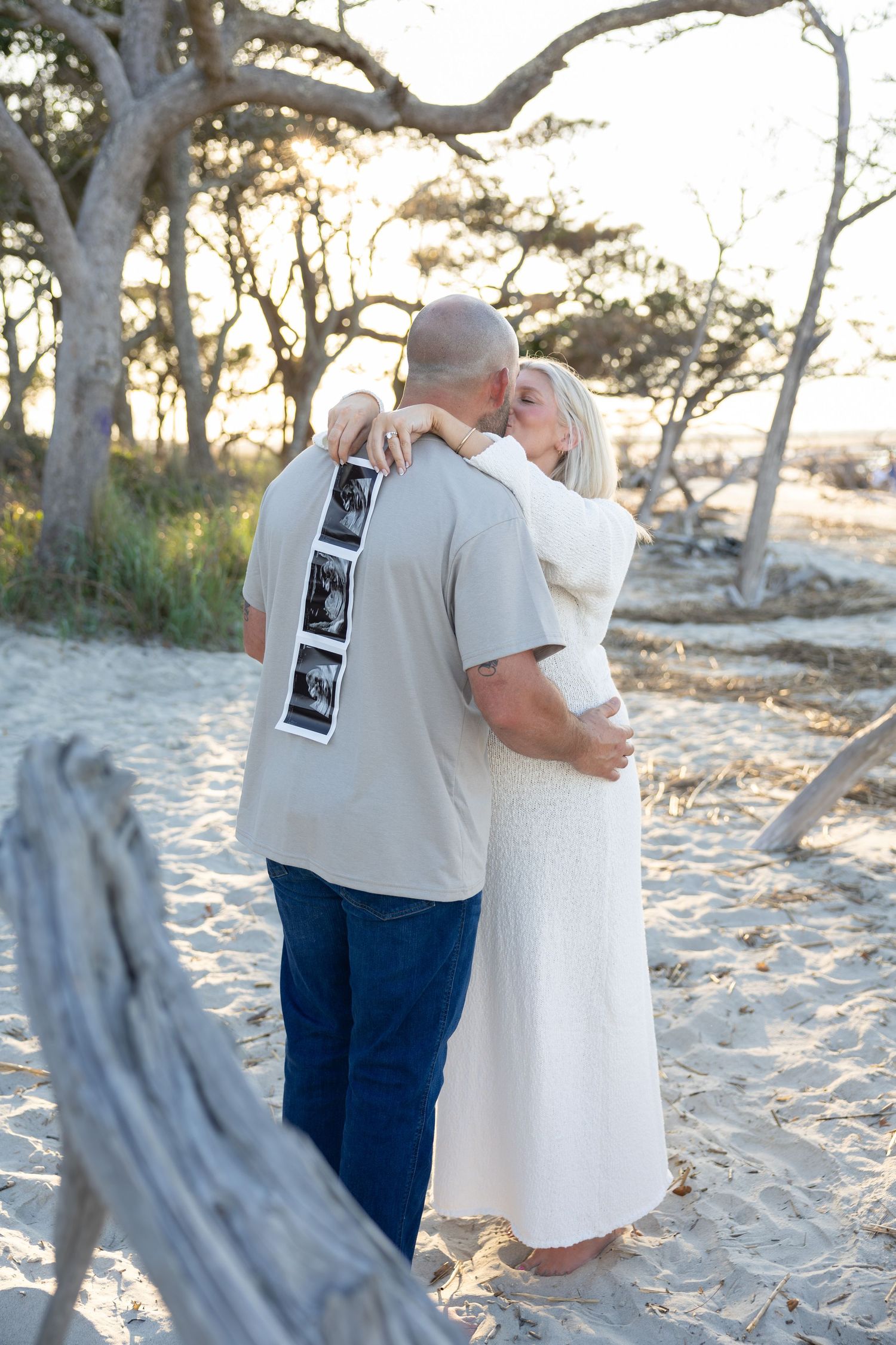Couple embracing on sandy beach holding ultrasound photos, announcing pregnancy near driftwood and trees at sunset.