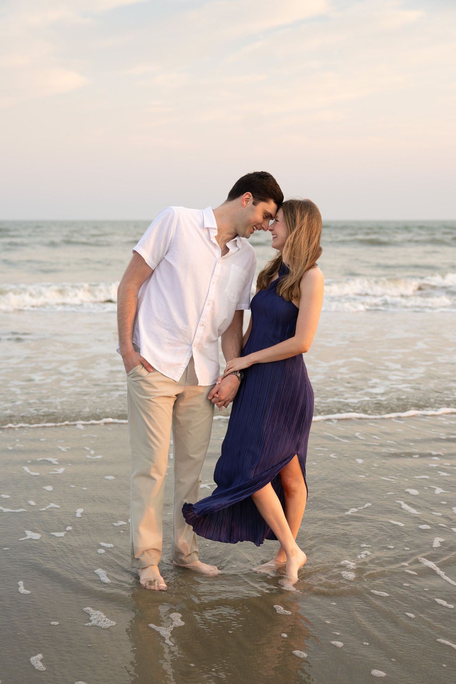 Romantic couple holding hands barefoot on a sandy beach at sunset, woman in blue dress, man in white shirt.