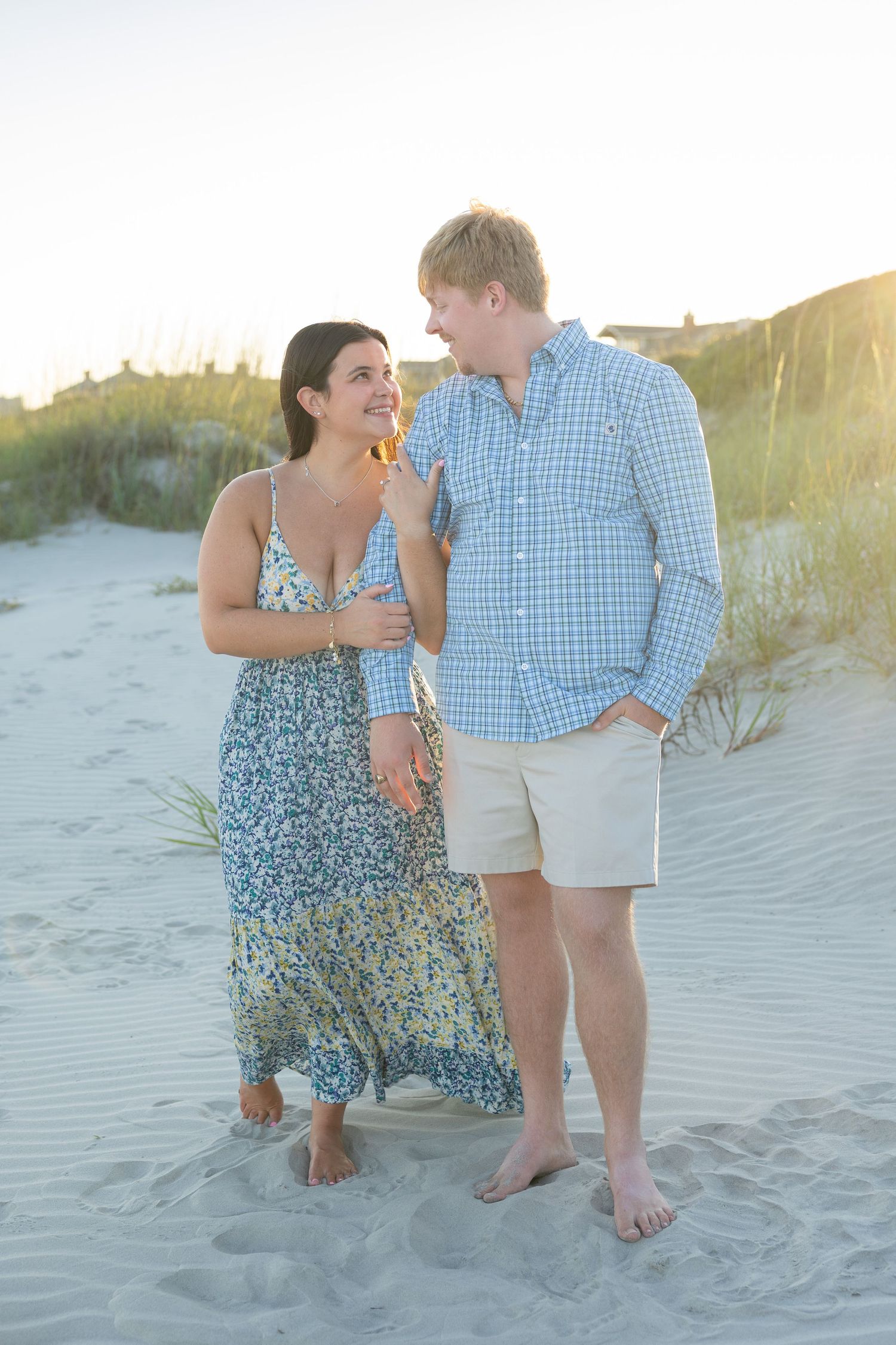 Couple smiling at each other on sandy beach at sunset, woman in floral maxi dress, man in blue plaid shirt and khaki shorts.