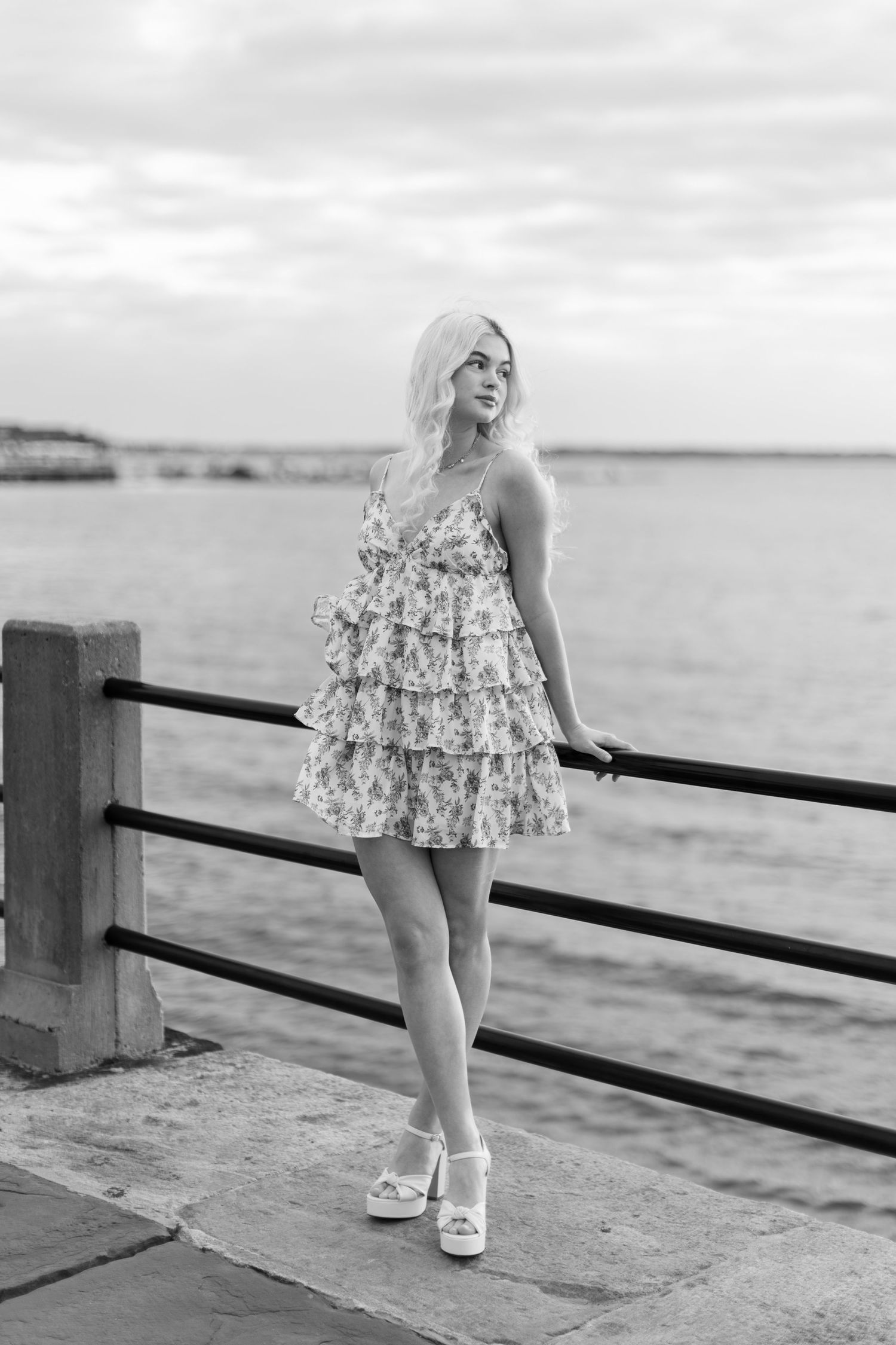 A black and white photo series shows a person in a floral dress posing along a seaside railing at sunset.