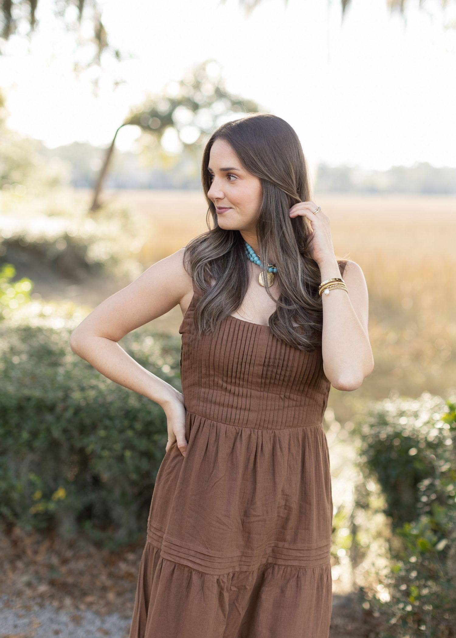 Woman in brown tiered maxi dress poses in outdoor setting with golden sunlight and natural scenery.