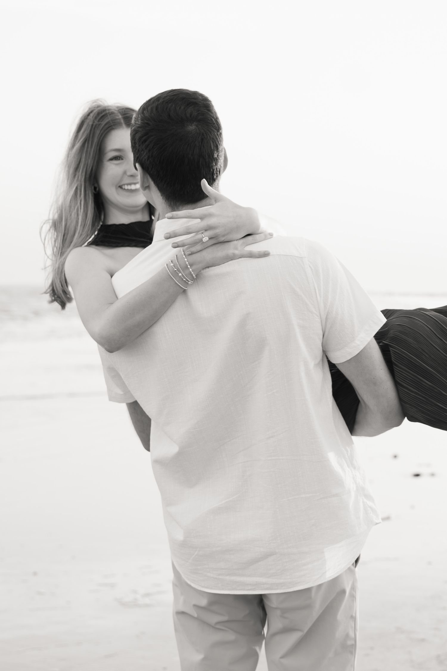 Black and white photo of a smiling woman embracing a man on a beach, wearing bracelets and a ring.