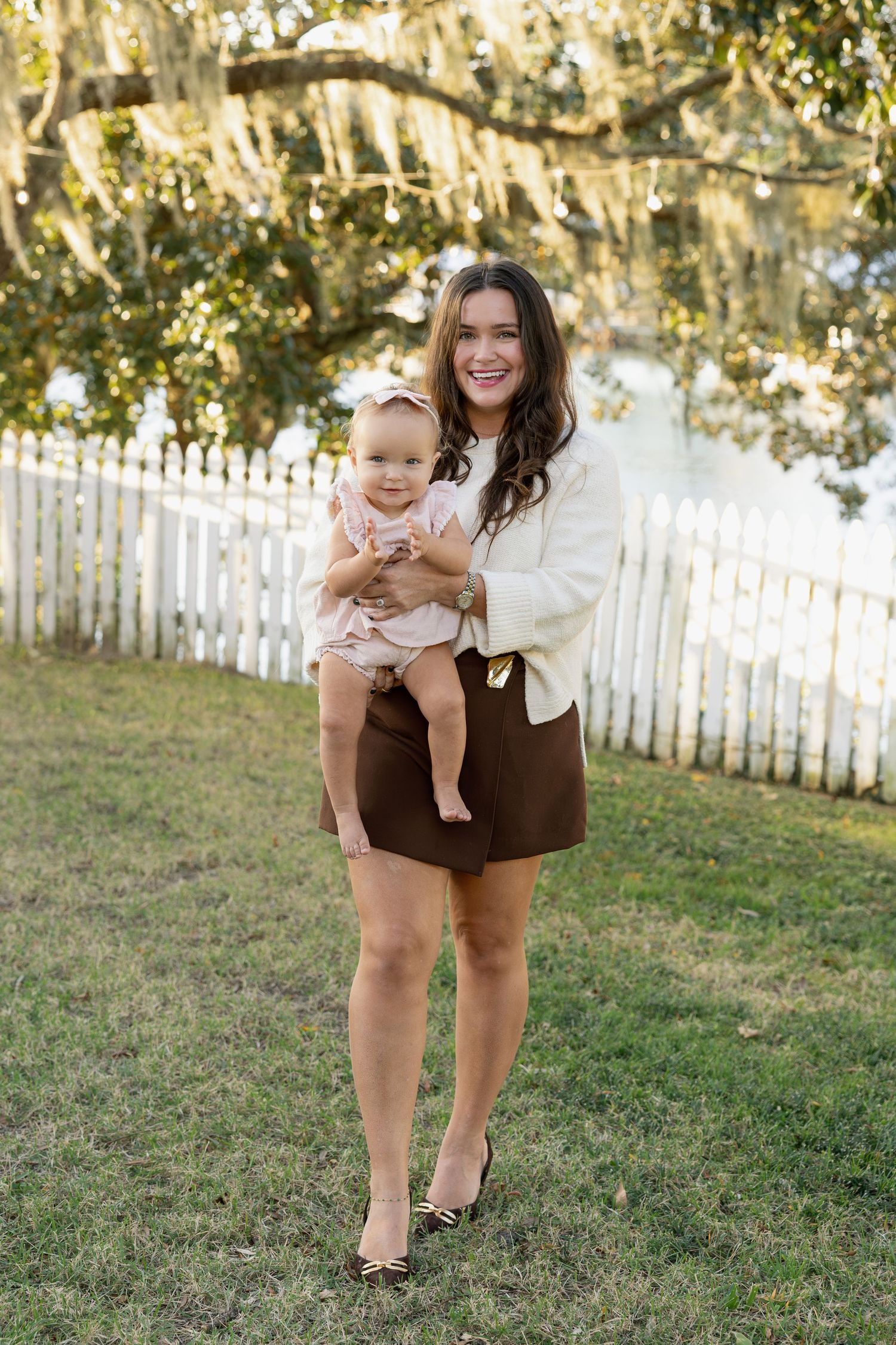 A parent and baby pose outdoors in front of a white picket fence with Spanish moss hanging from trees in the background.