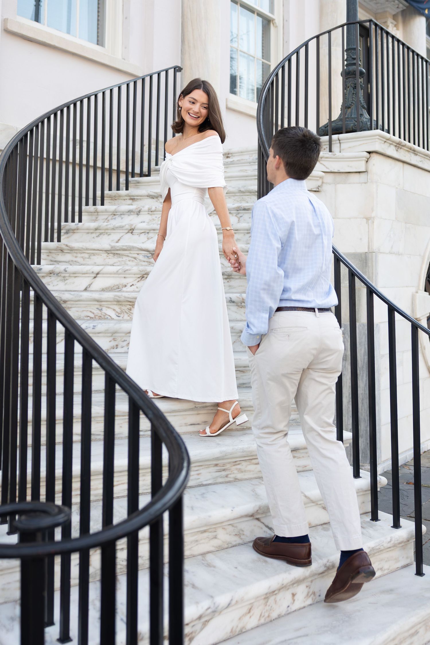 Couple holding hands on marble spiral staircase, woman in white off-shoulder dress, man in light blue shirt and khakis.