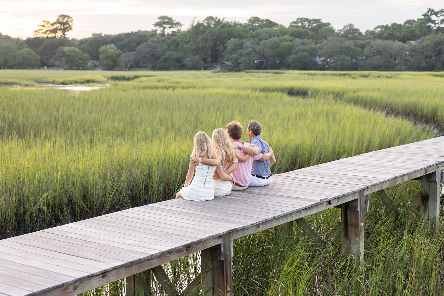 Family sitting together on a wooden dock overlooking a lush green marsh at sunset.