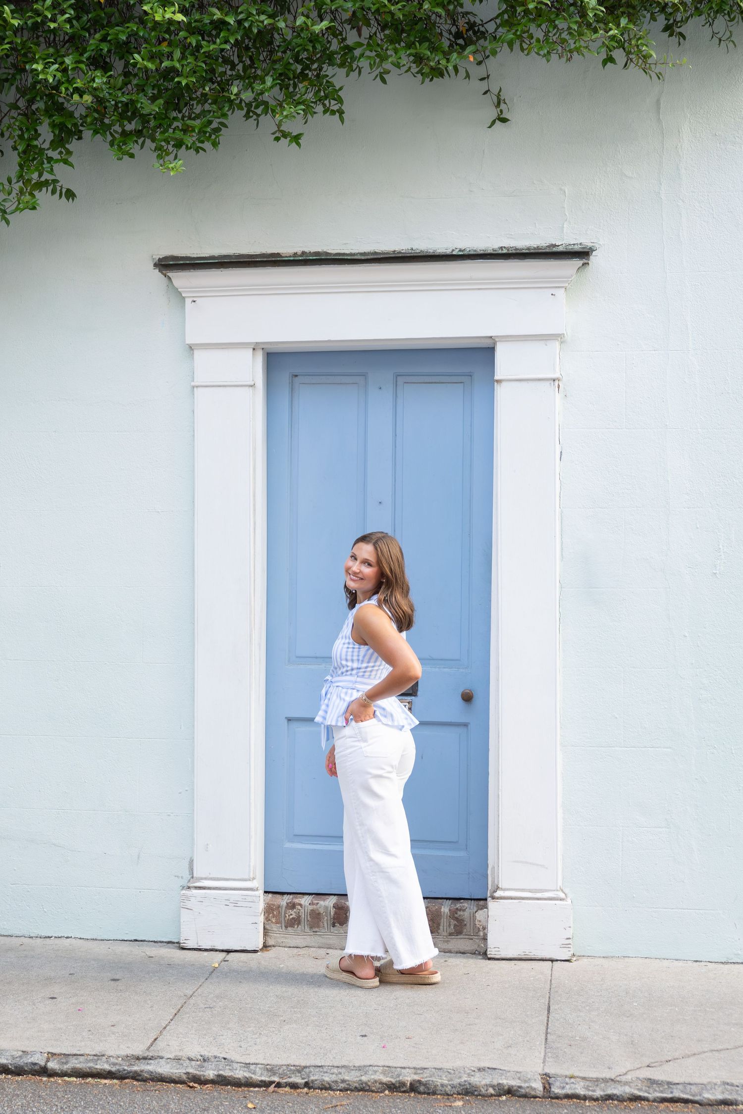 Young woman in white wide-leg pants and blue top posing by a charming blue door on a pastel wall.