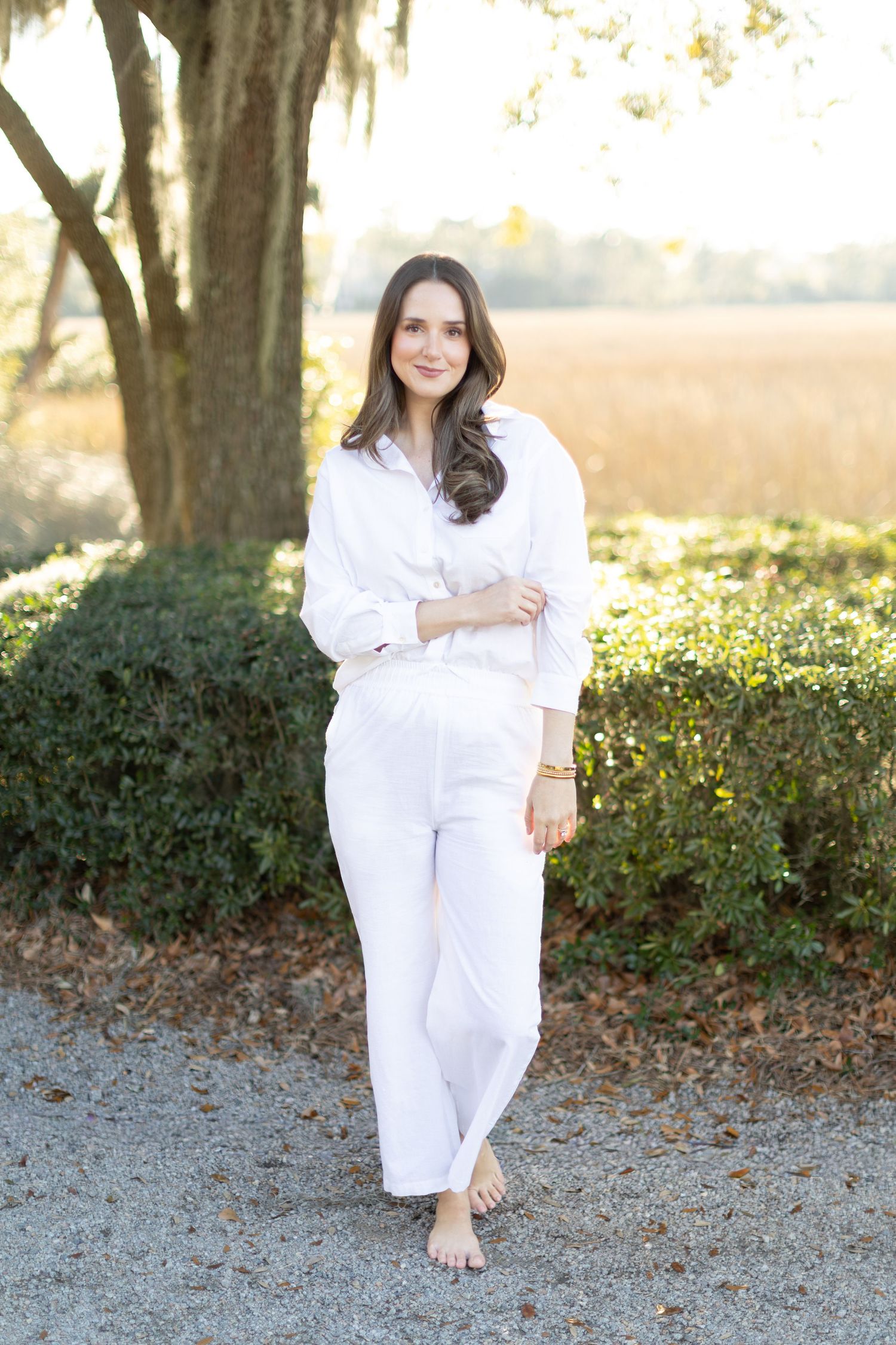 A person in an all-white outfit stands barefoot on a gravel path with sunlit trees and foliage in the background.