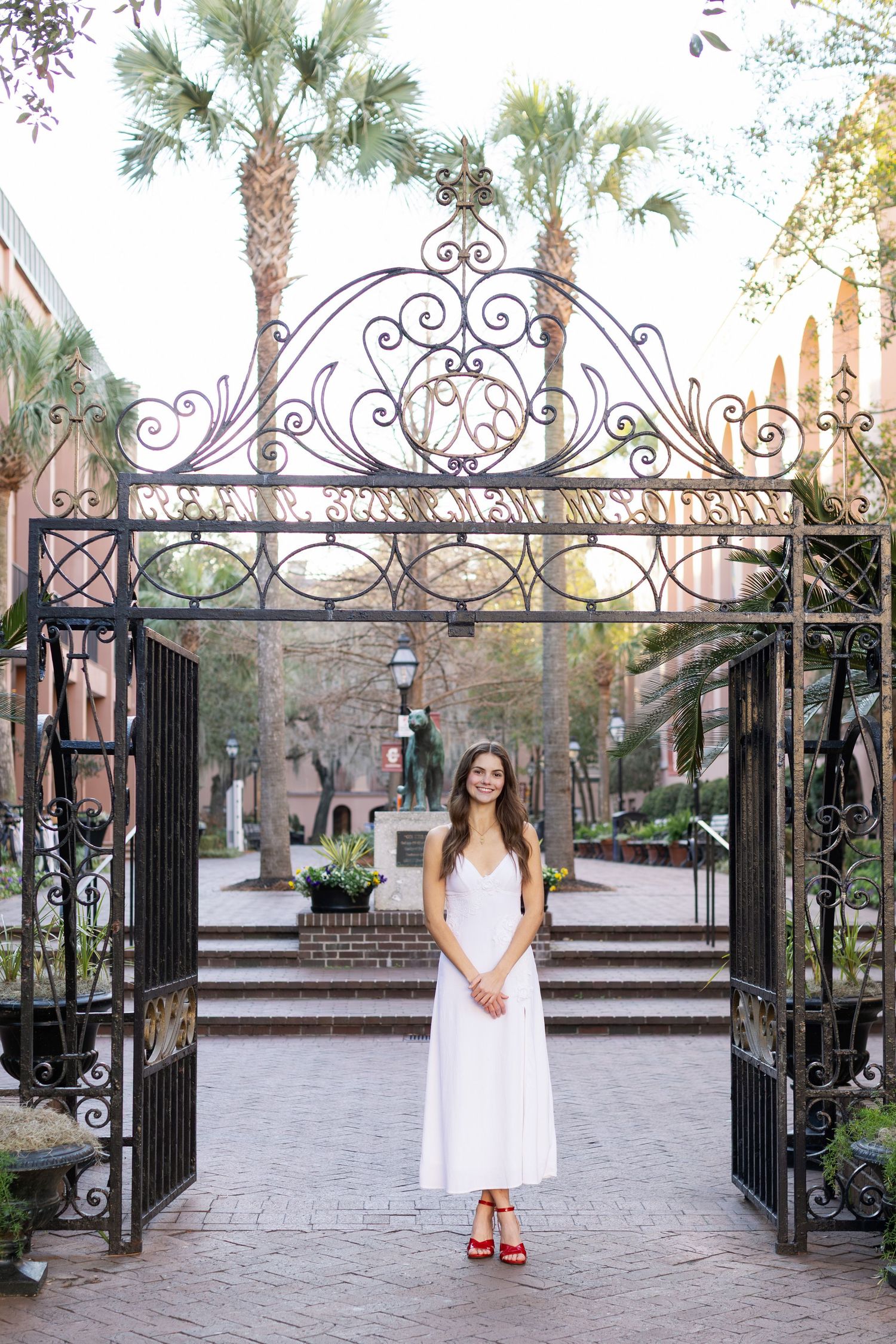 A series of elegant wrought iron gates frame a brick paved courtyard with palm trees and landscaping in the background.