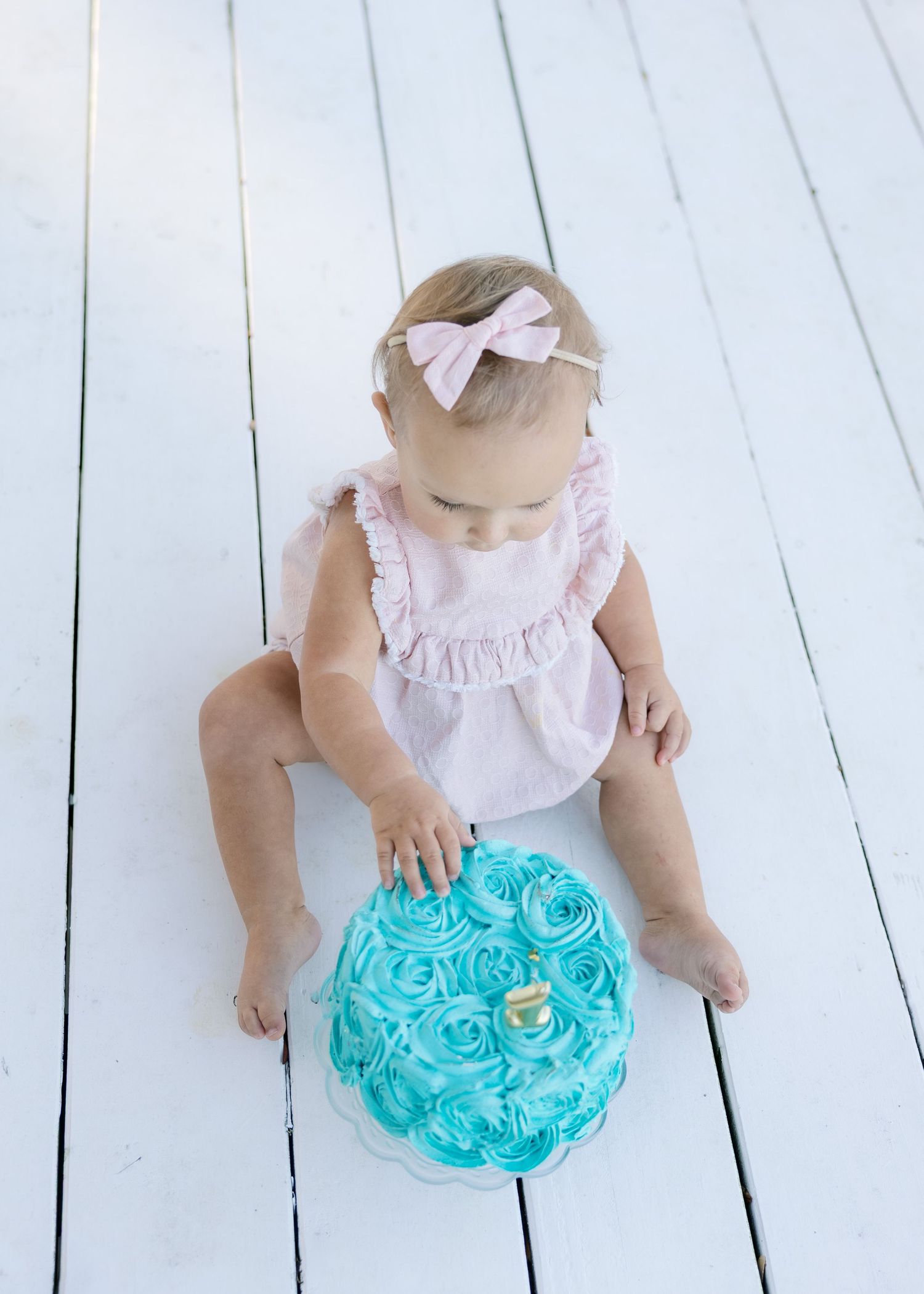 Baby in pink ruffled top and turquoise tutu skirt sits on white wooden floor during photo session.