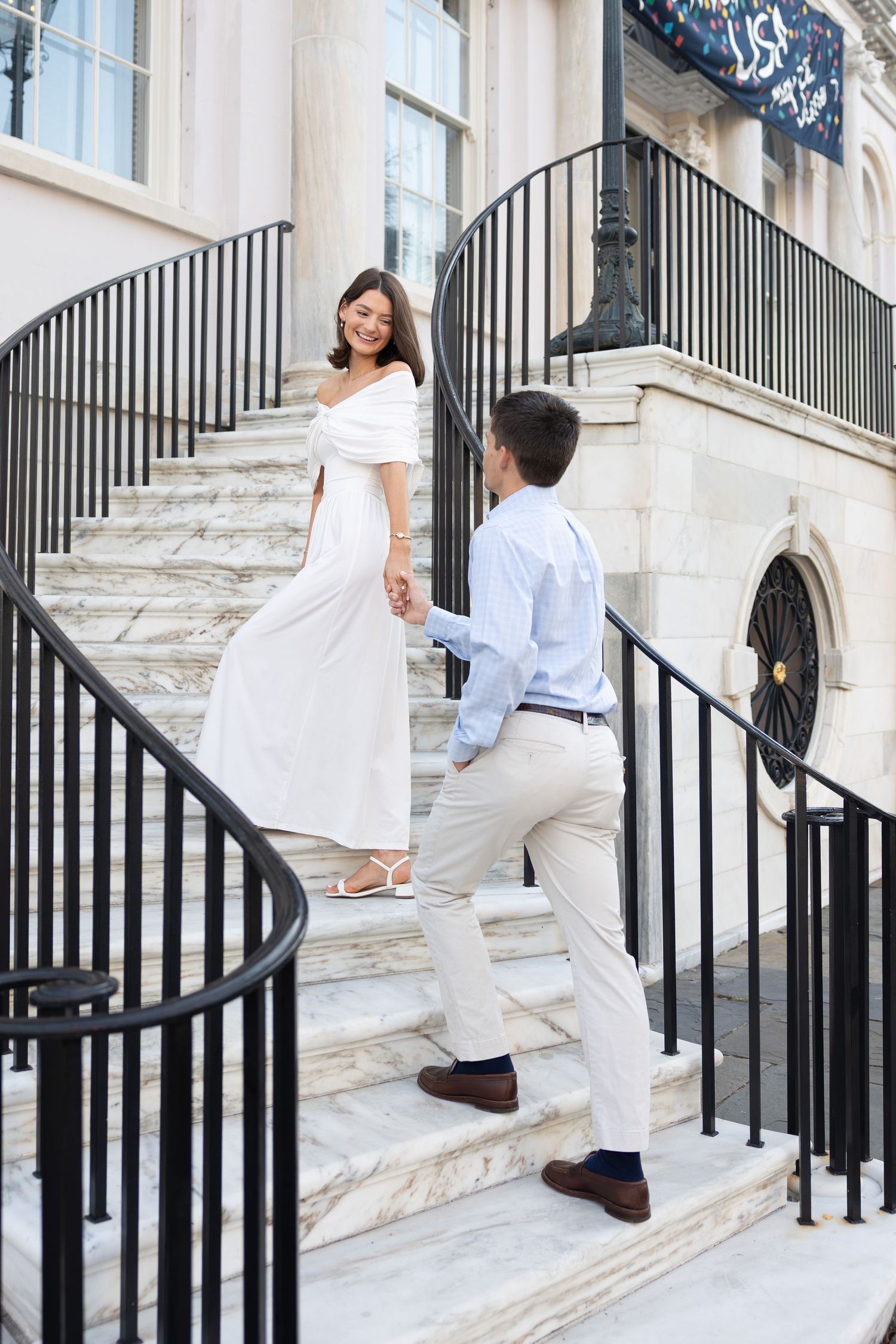 Couple holding hands on elegant marble staircase, woman in white dress looking back, man in light blue shirt.