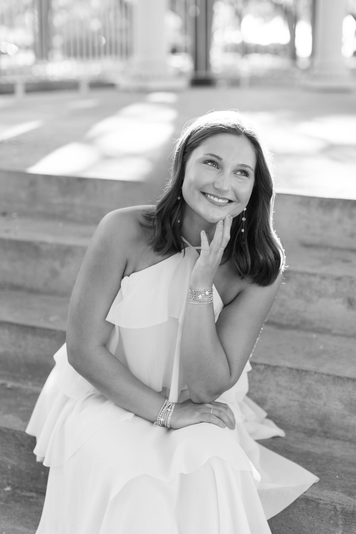 Black and white photo of a smiling young woman in a strapless white dress sitting on steps.