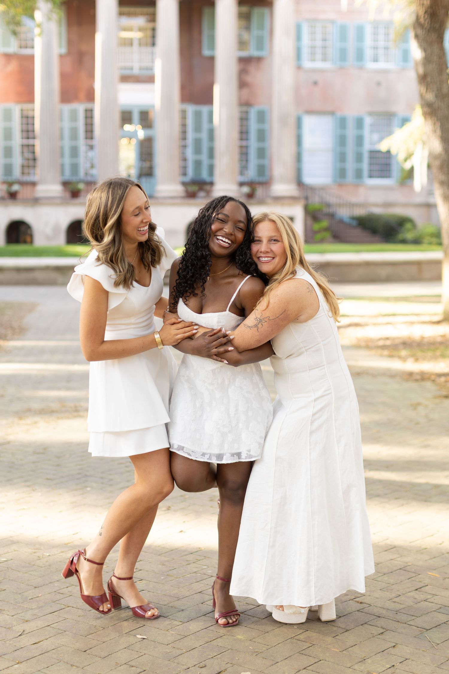Three women in white dresses hugging and smiling outdoors near a brick building on a sunny day.
