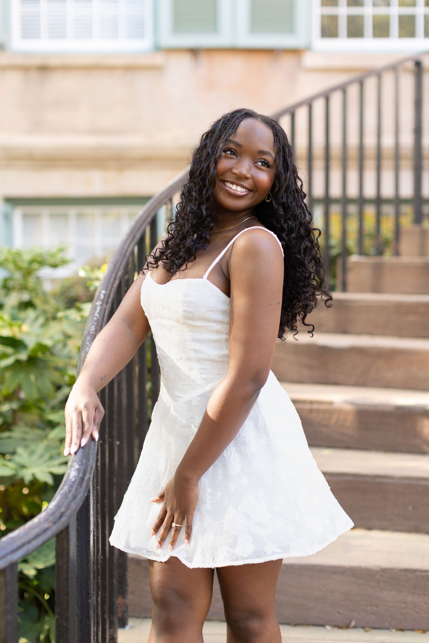 Young Black woman in white mini dress smiling on outdoor steps with lush greenery background.