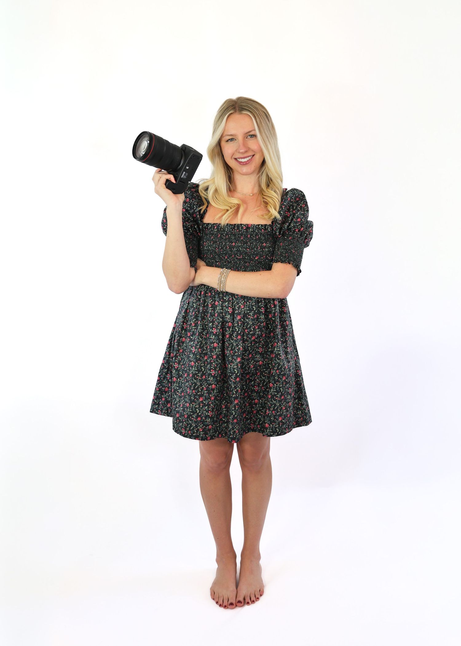 Professional photographer in floral dress holding camera lens against white backdrop.