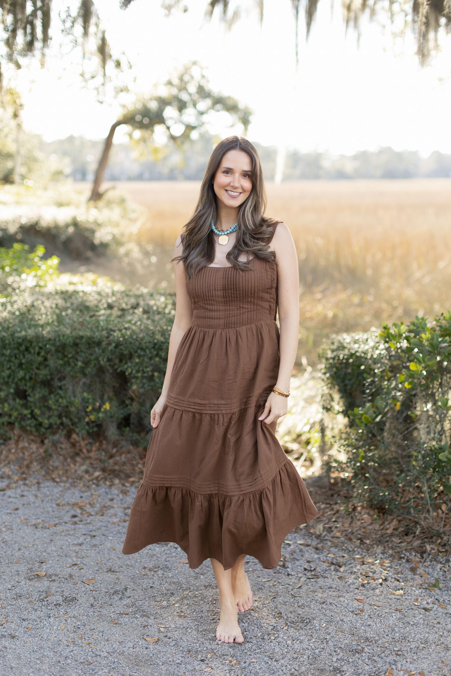 A brown tiered cotton dress is showcased in a series of outdoor photos with greenery and sunlight in the background.