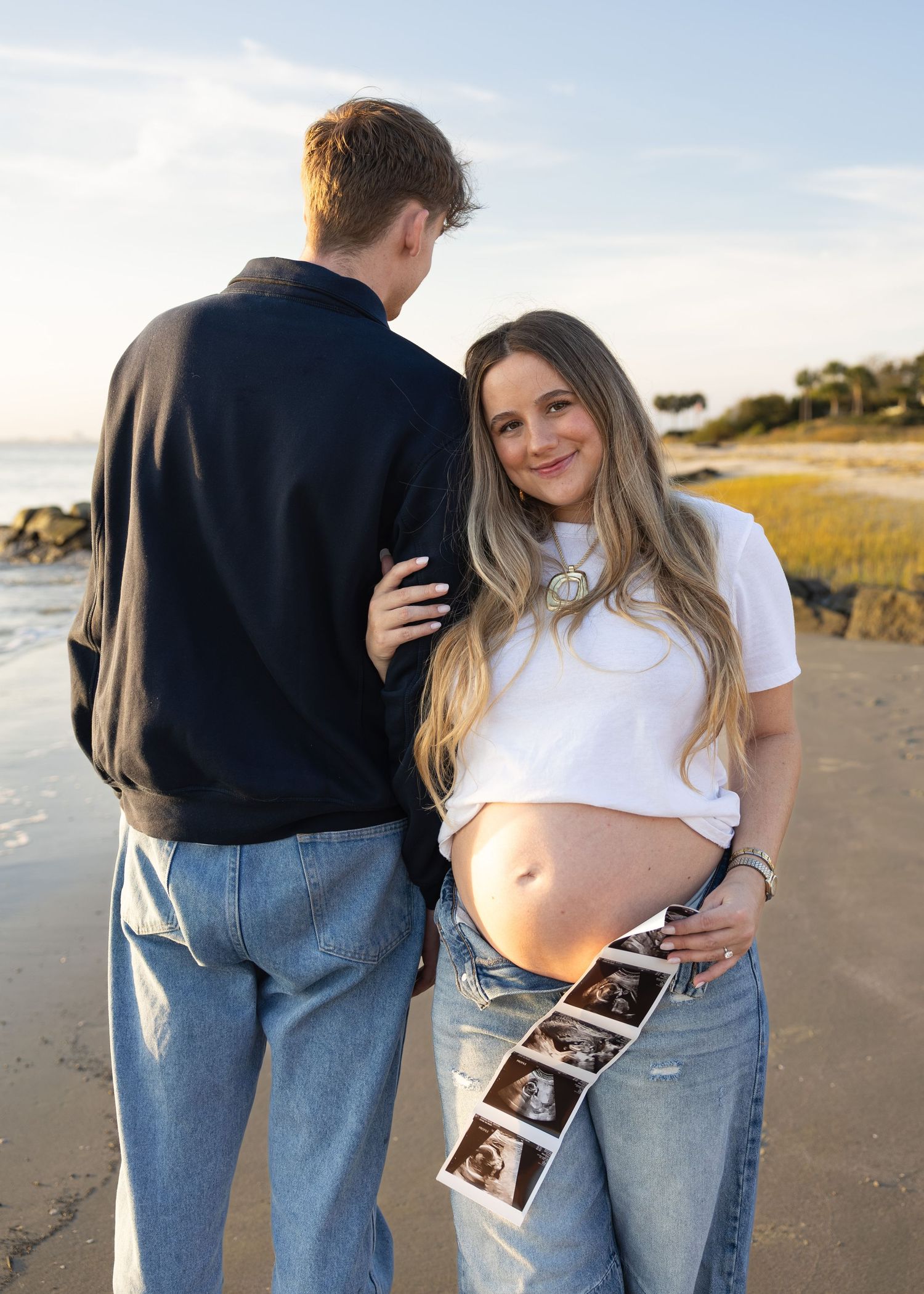 A couple poses with ultrasound photos on a beach during sunset for a pregnancy announcement photoshoot.