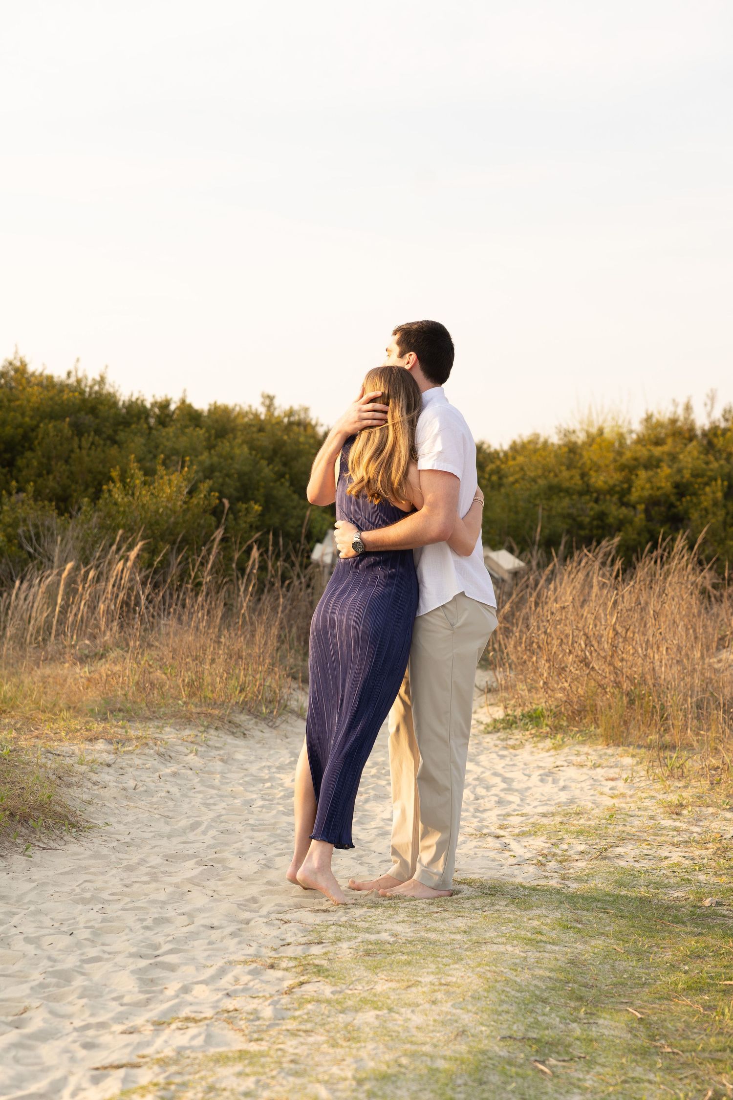 Couple embracing on sandy beach path surrounded by golden grass and green shrubs at sunset.