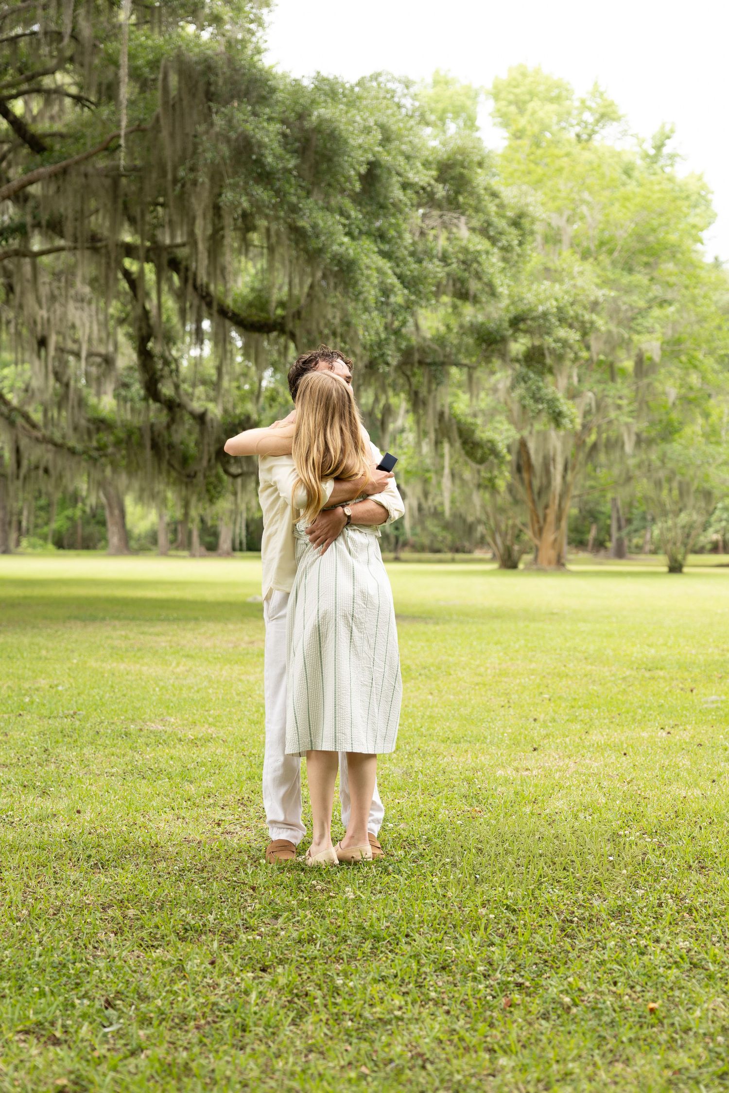 Couple embracing barefoot on lush green grass beneath sprawling moss-draped oak trees in a romantic outdoor setting.