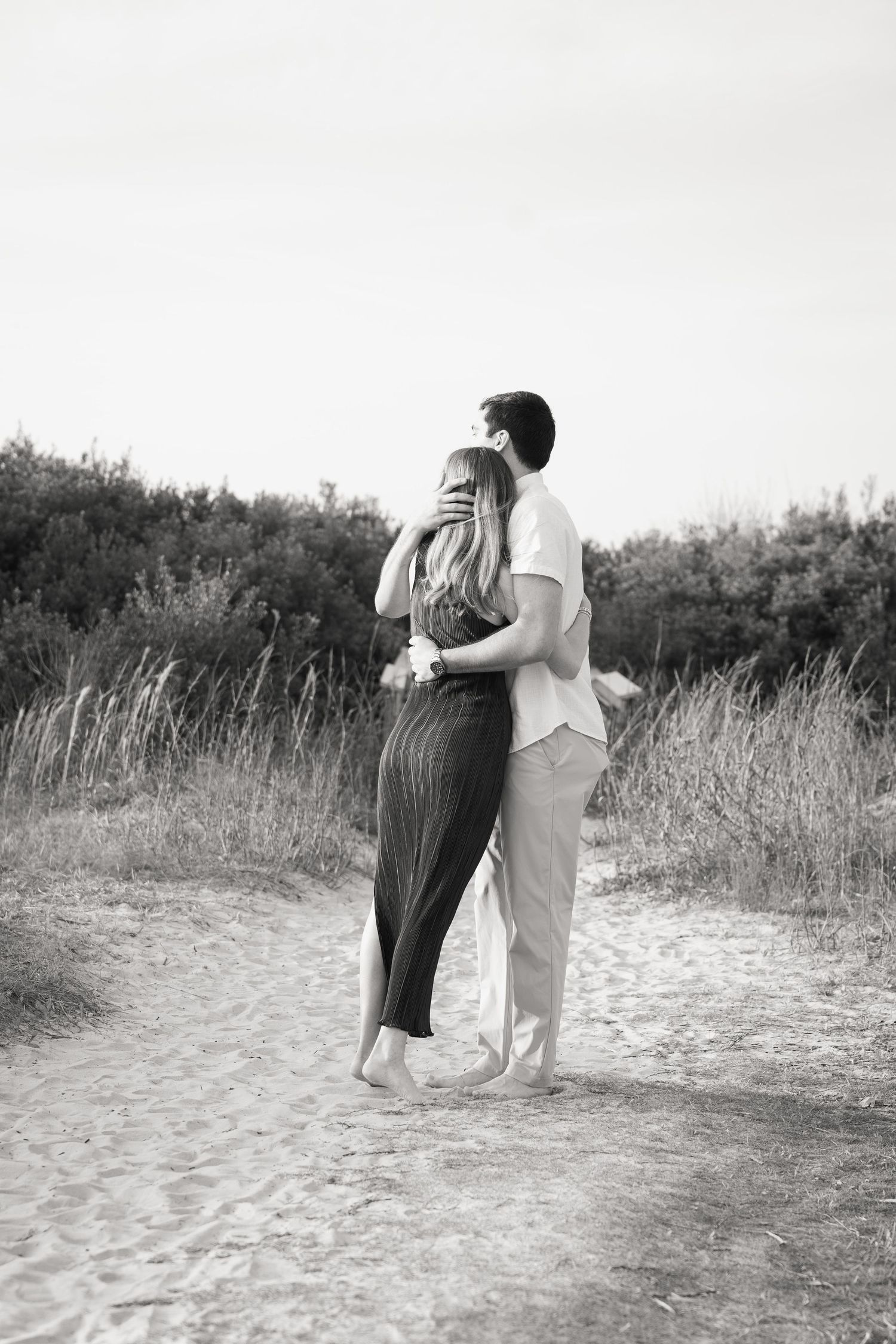 A couple embraces while walking along a sandy path through tall grasses in a series of black and white photographs.