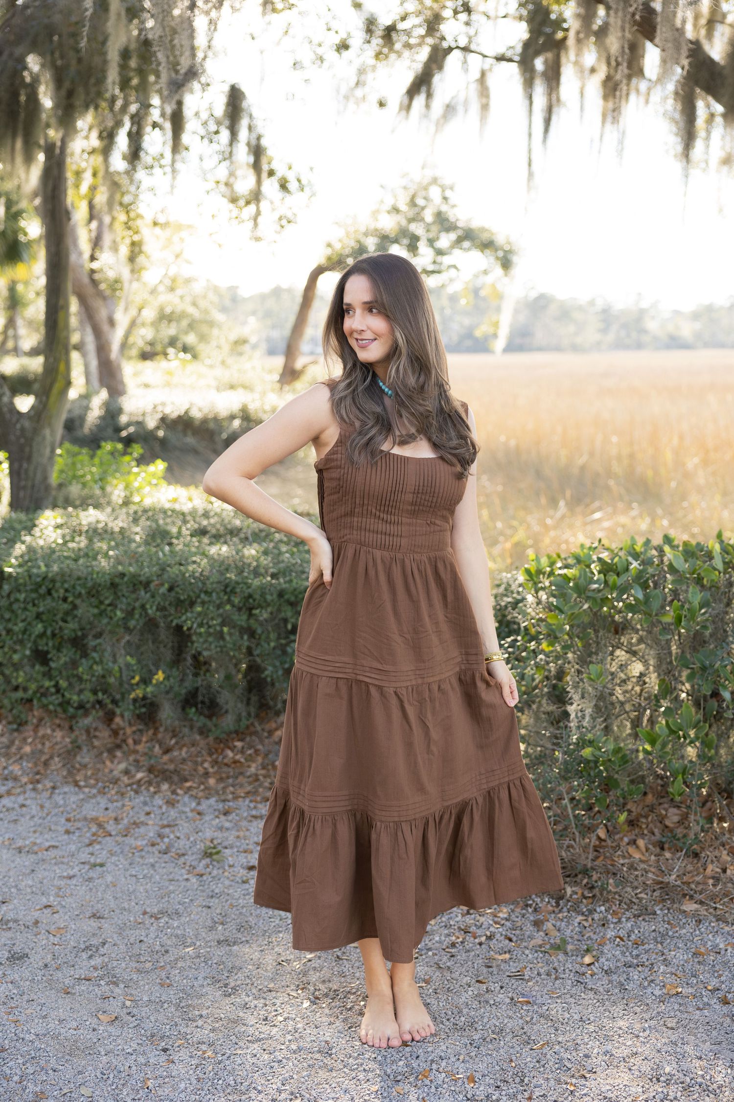 A tiered brown cotton sundress is modeled in an outdoor setting with Spanish moss and marsh grass in the background.