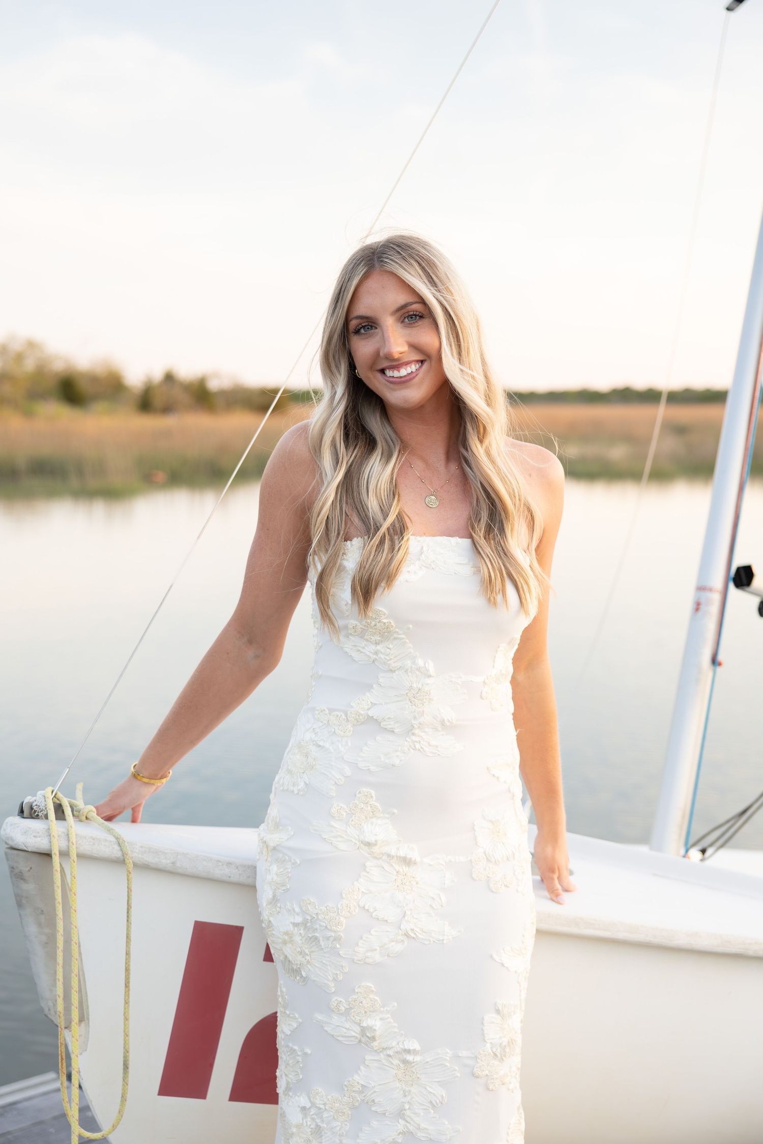 Smiling blonde woman in white floral strapless gown posing on a sailboat deck with calm water in background.
