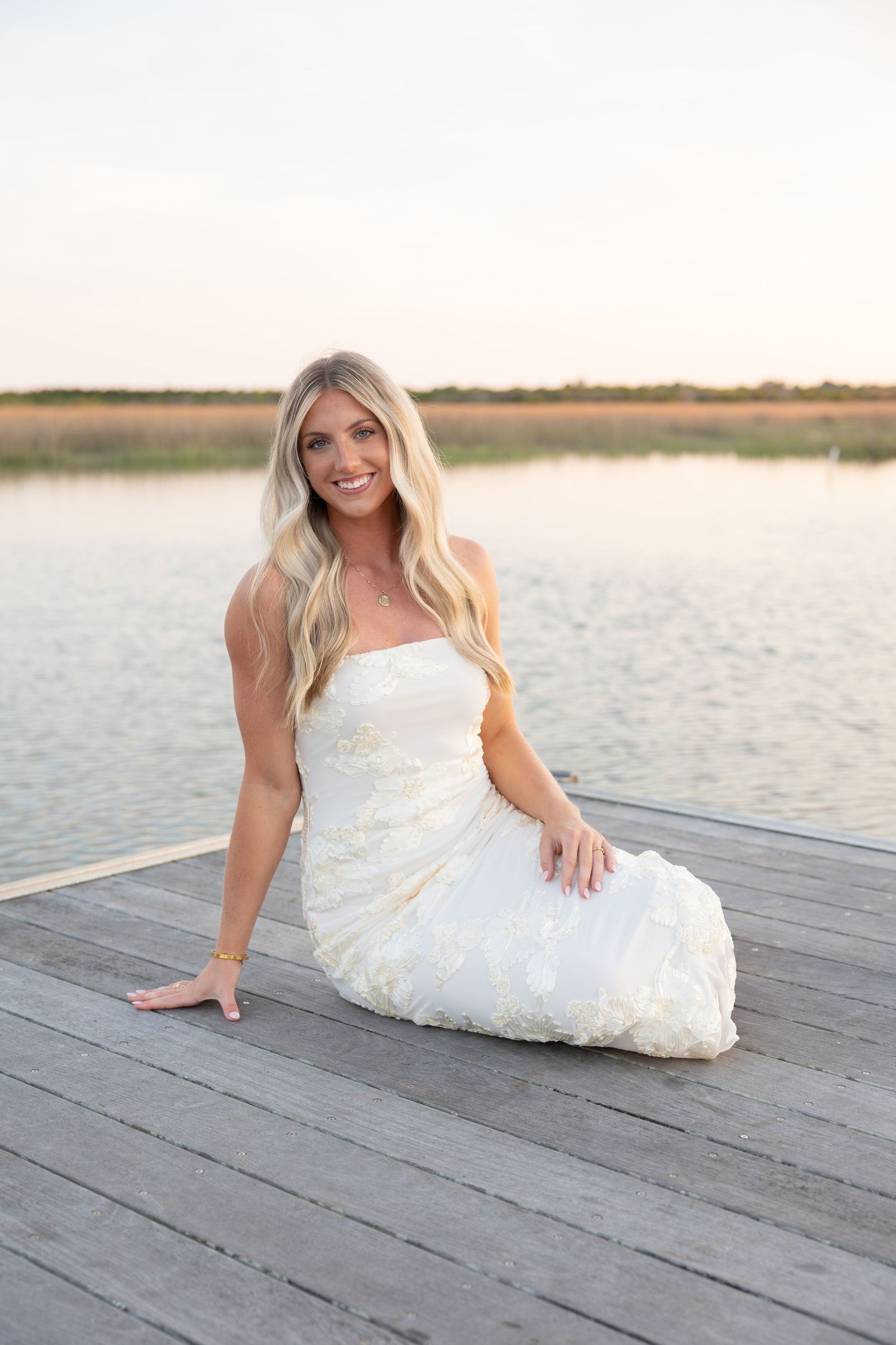 Smiling blonde woman in white strapless gown sitting on wooden dock by calm lake at sunset.