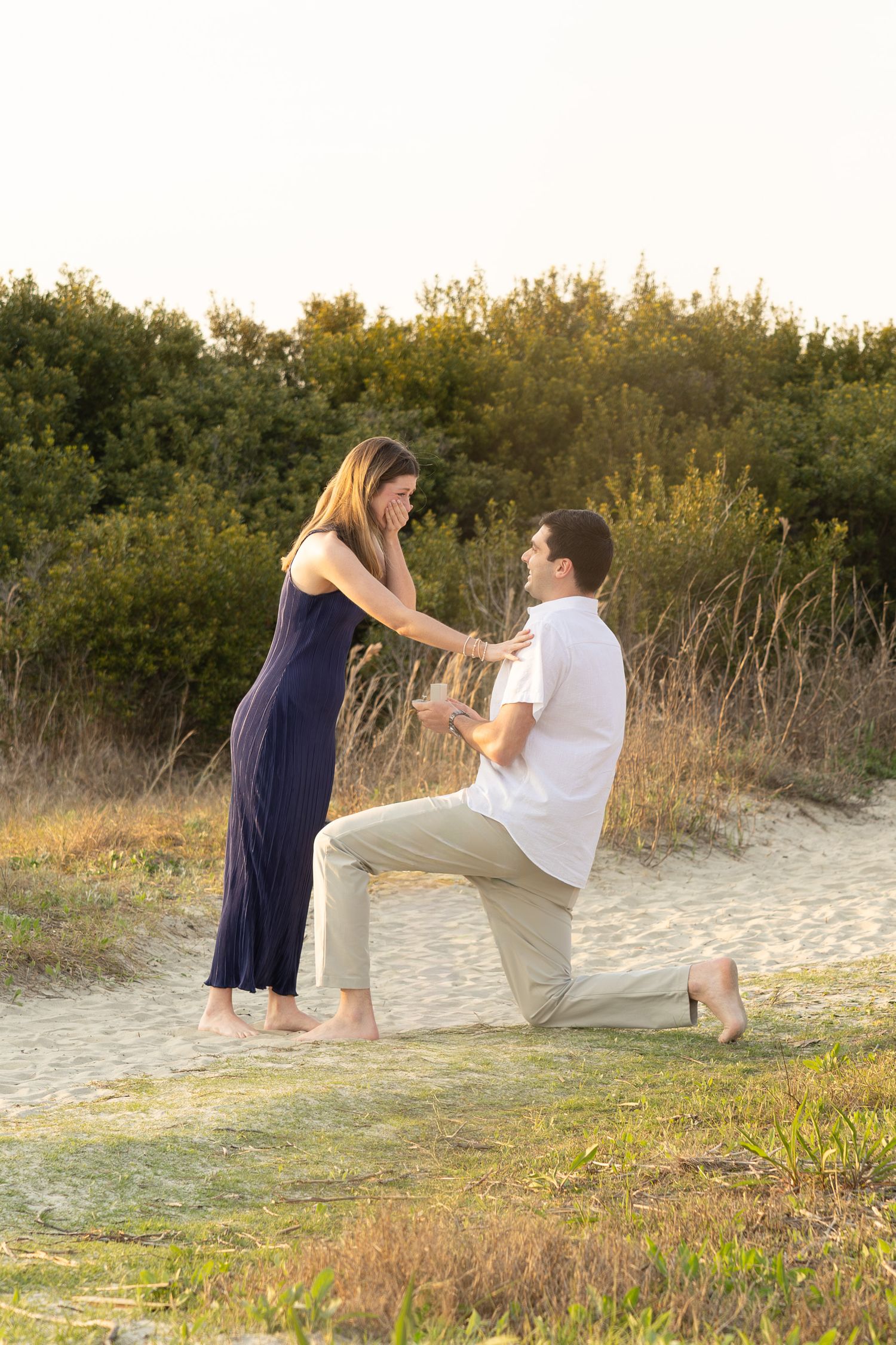 A couple proposing on a sandy beach path with green foliage and golden sunlight in the background.