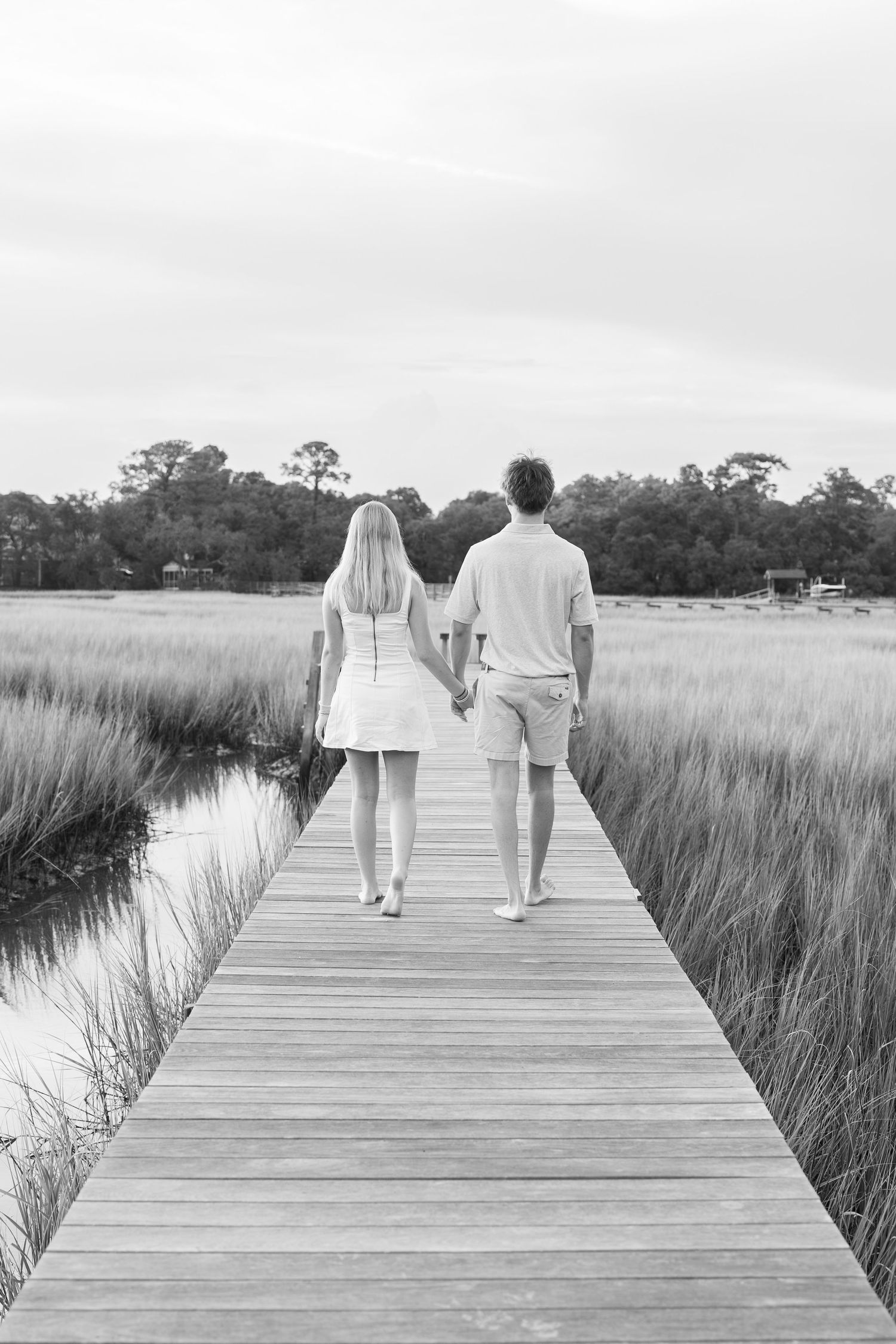 Couple holding hands walking on wooden boardwalk through marsh grass in black and white photography.