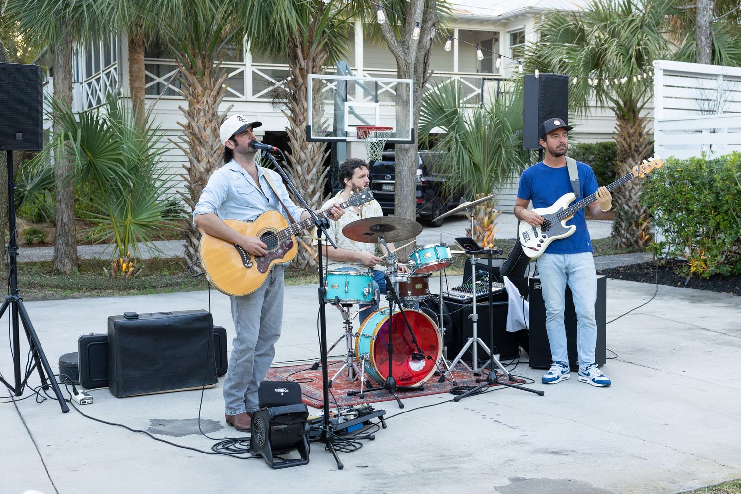 Musicians perform with acoustic guitars and other instruments in an outdoor courtyard area with palm trees.