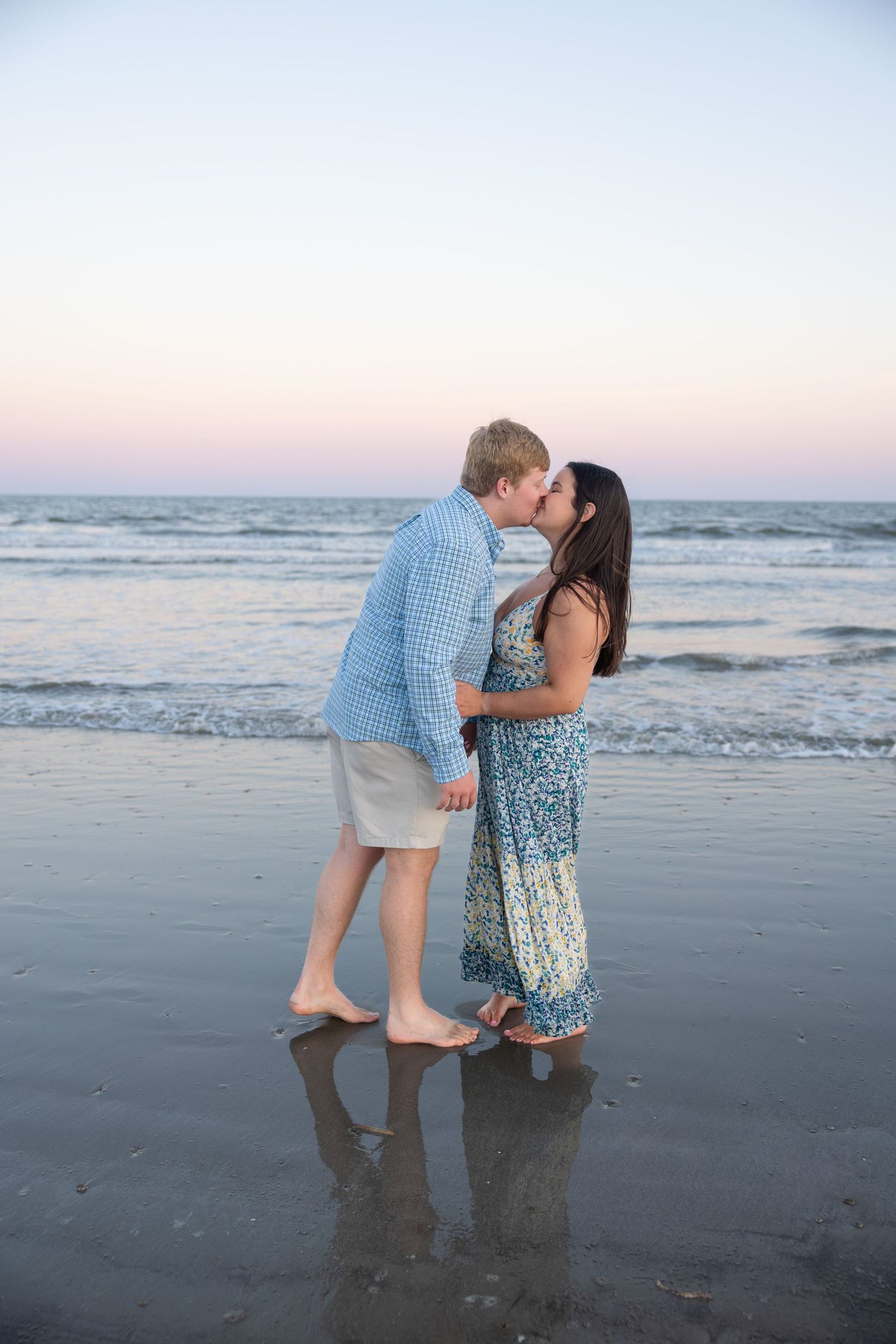 Couple sharing a romantic kiss on a sandy beach at sunset, waves and pastel sky in the background.