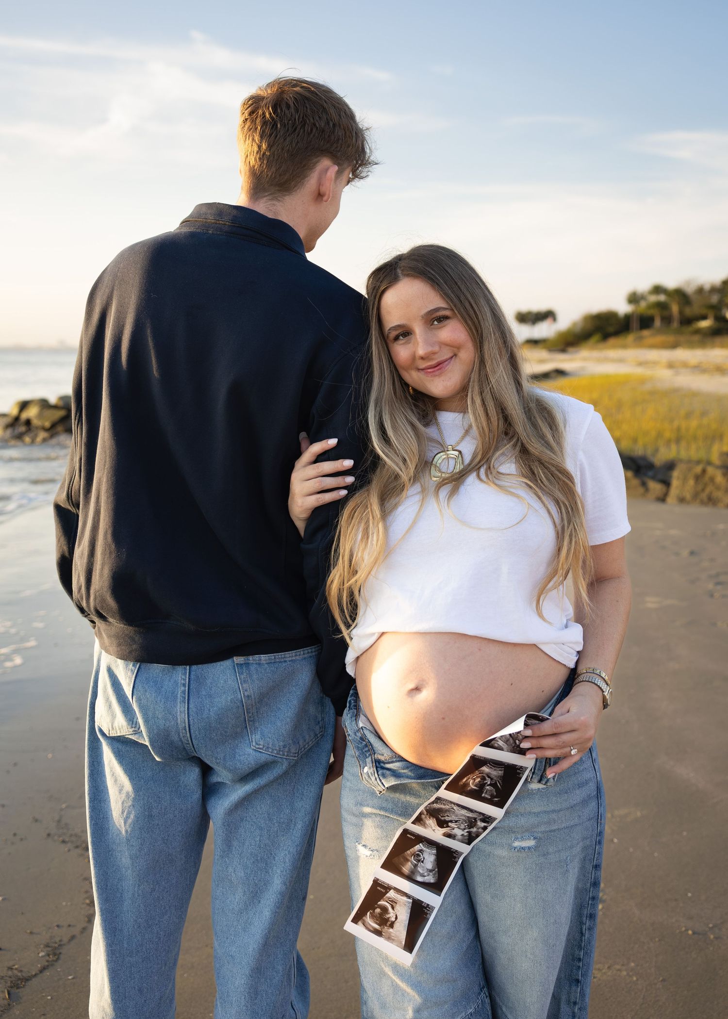 Pregnant woman smiling on beach holding ultrasound photos beside partner, maternity announcement photo at sunset.