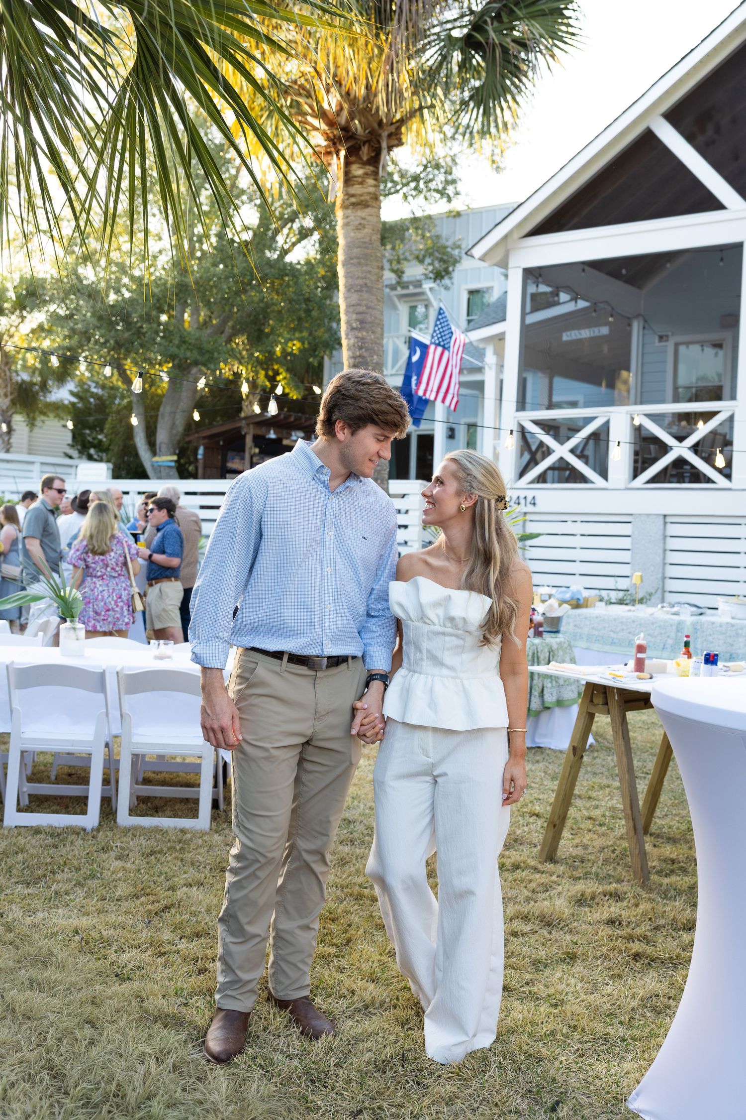 Couple holding hands at outdoor garden party near palm tree with American flag and white house backdrop.