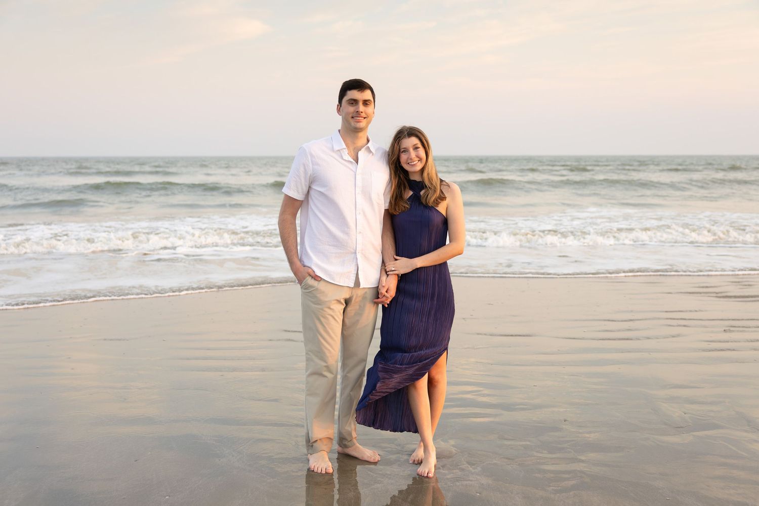 Couple posing together on a sandy beach at sunset, man in white shirt and woman in navy dress.