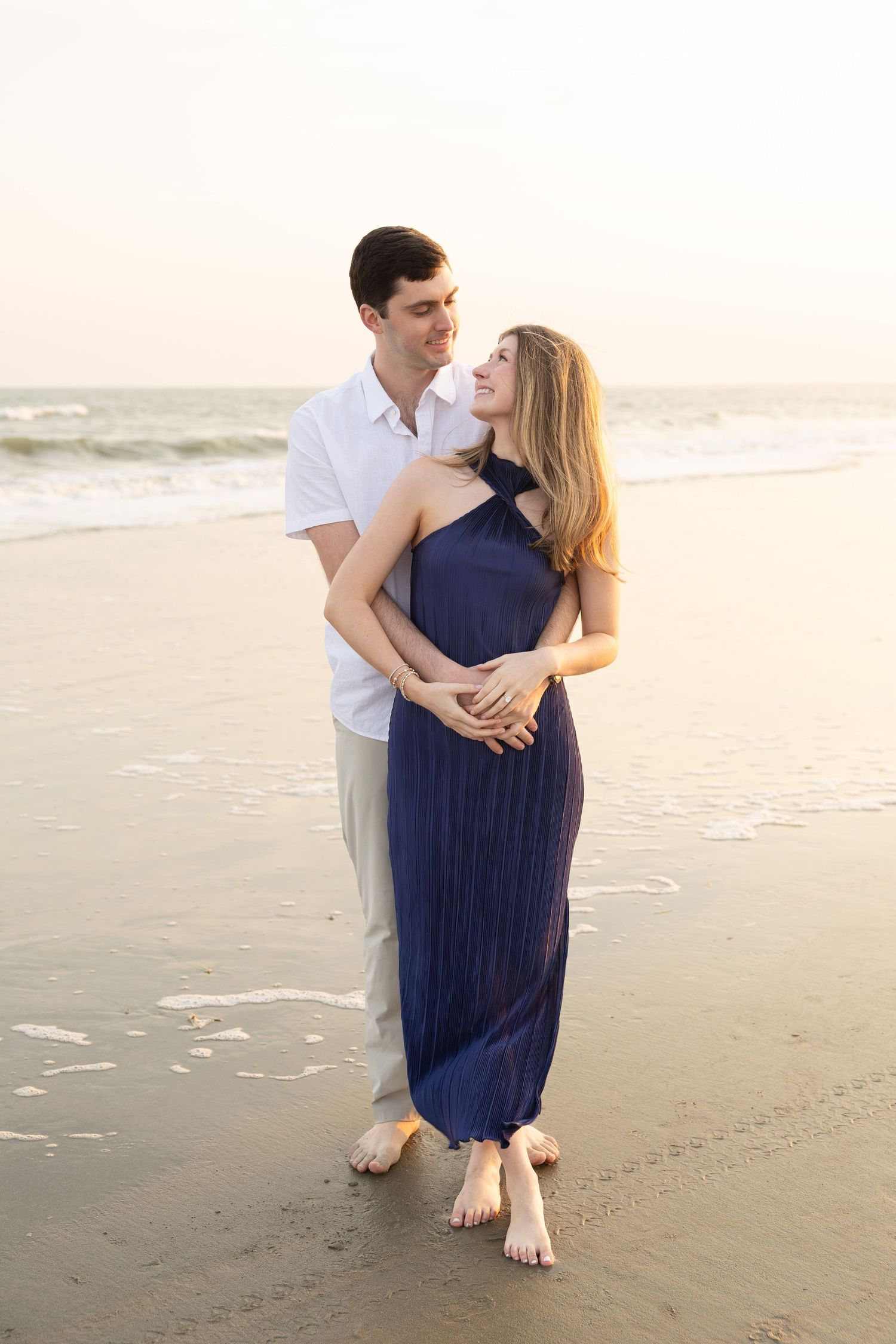 A couple embraces barefoot on a beach at sunset, woman in navy dress smiling up at man in white shirt.
