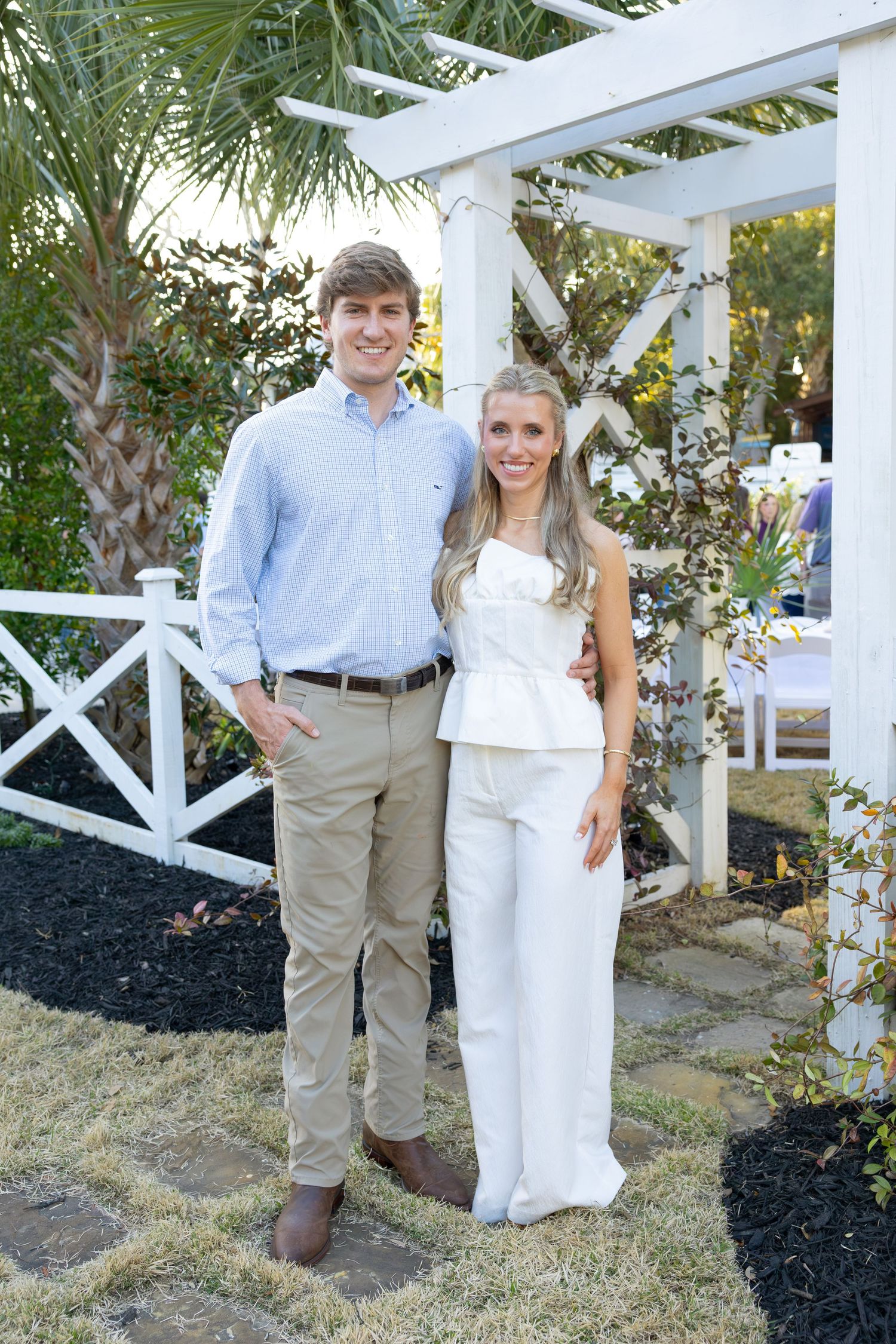 A couple poses together in front of a white pergola and palm trees wearing coordinated light-colored outfits.