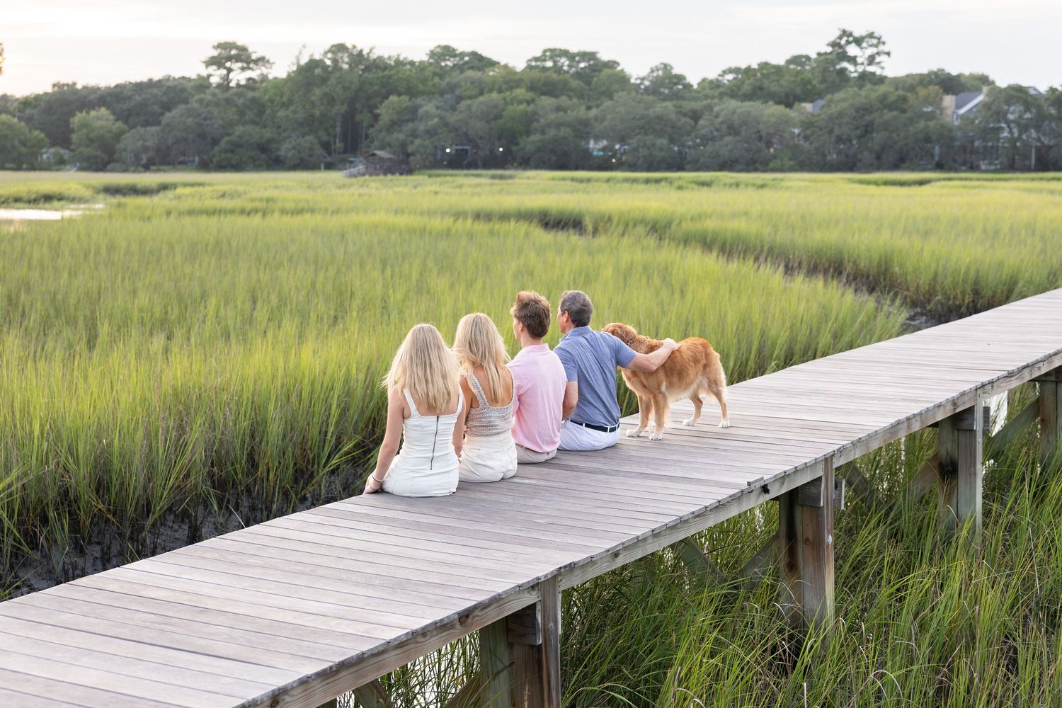 Family with golden retriever dog sitting on wooden dock overlooking lush green marsh wetlands at dusk.