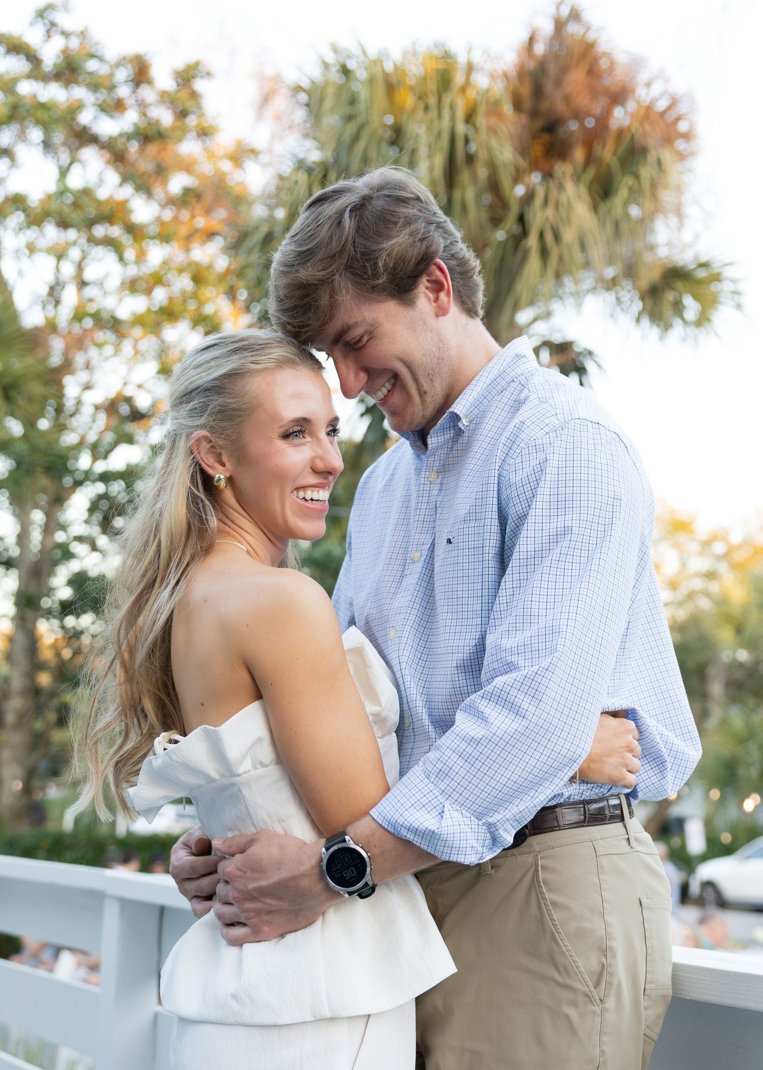 A smiling couple embraces outdoors near palm trees, woman in white strapless dress, man in light blue plaid shirt and khakis.