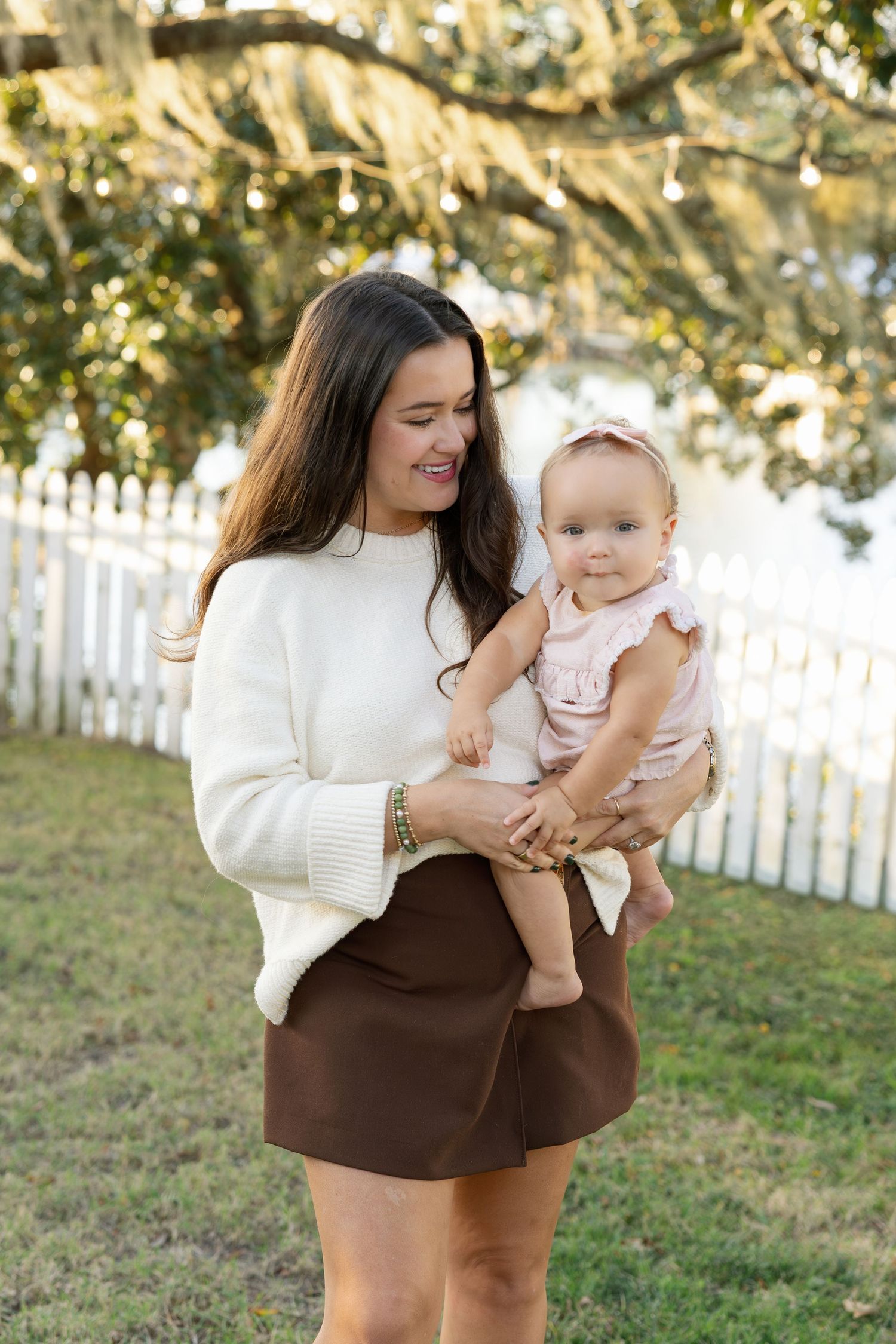 A parent holds a baby in a white sweater and brown skirt against a backdrop of trees and white picket fence.