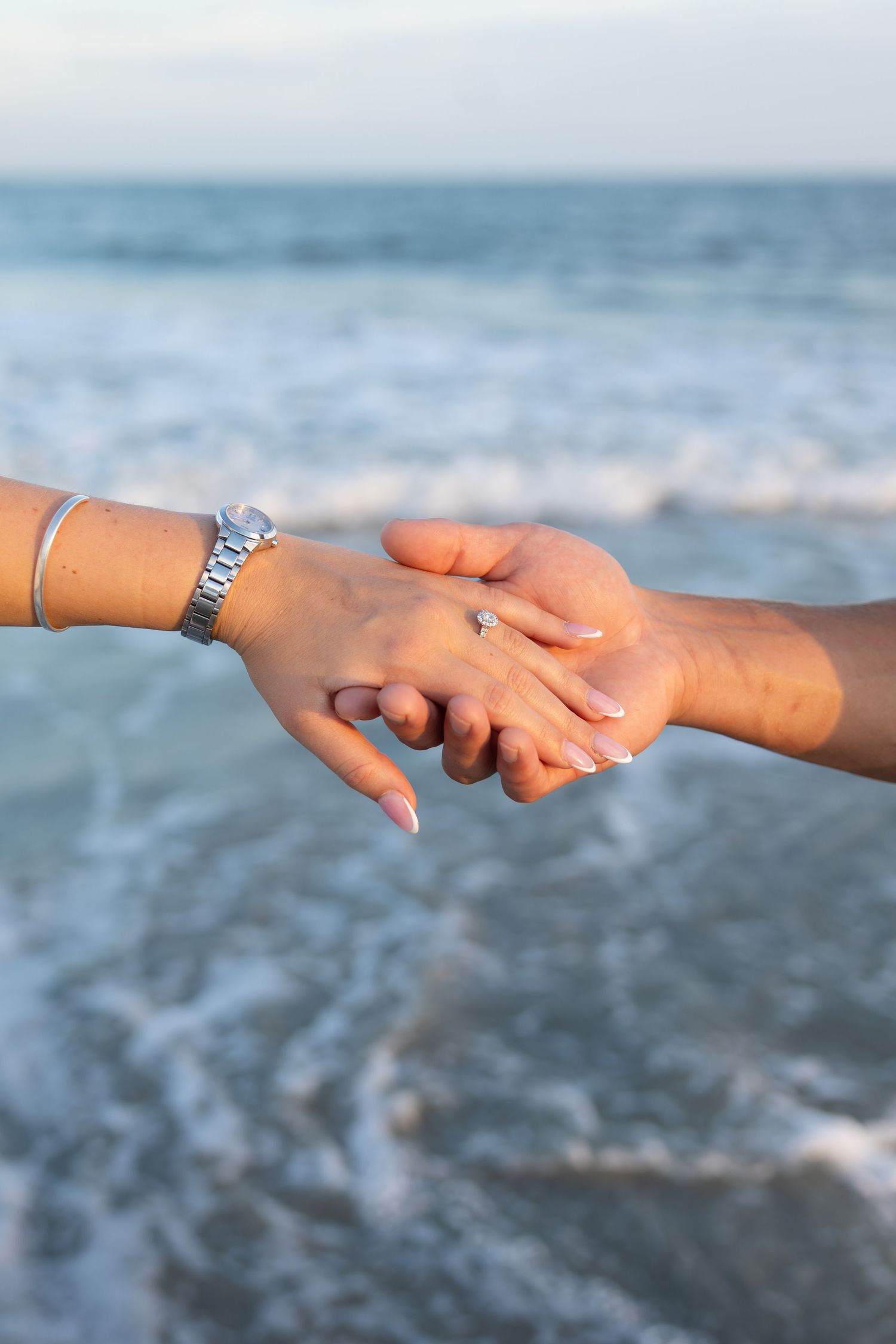 Two hands holding at the beach with an engagement ring and silver bracelet visible, ocean waves in the background.