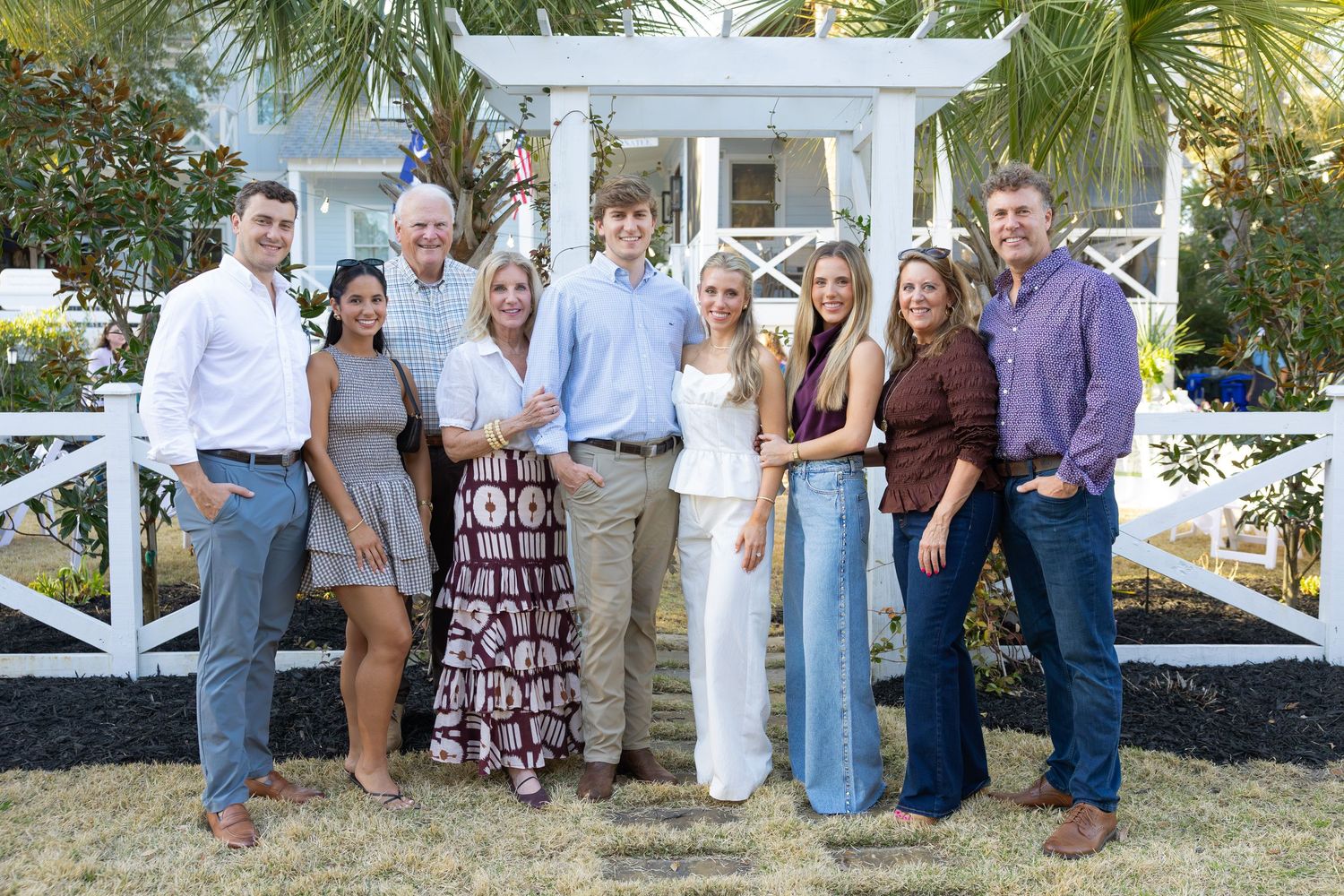 Group of people gathered outdoors near white fencing and palm trees at a casual event.