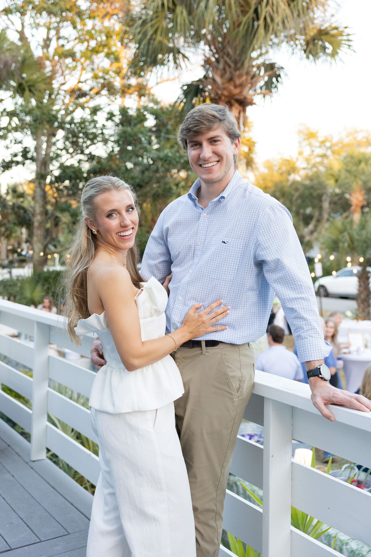 Romantic couple poses along white railing at sunset with palm trees and natural lighting in background.