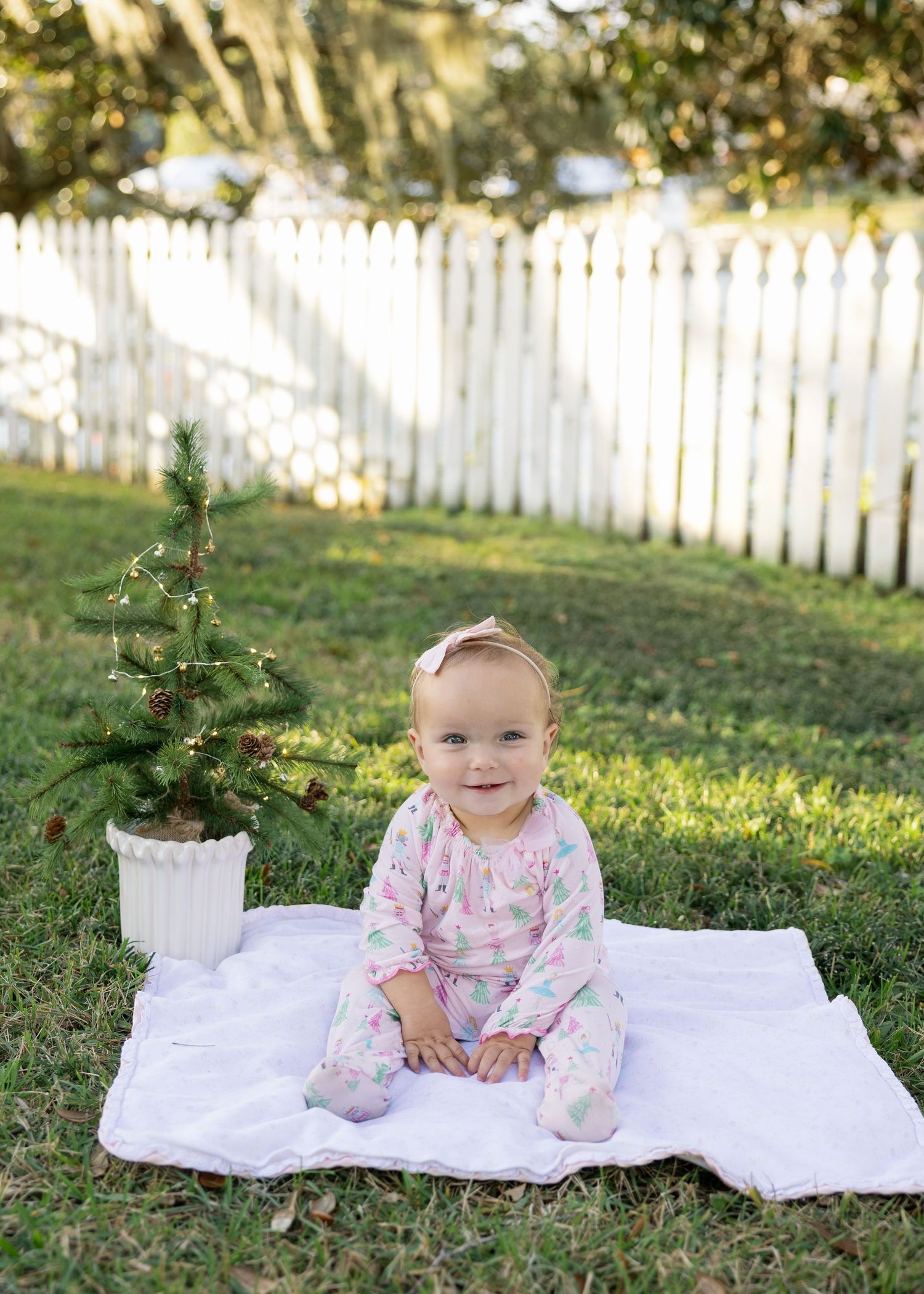 Baby in floral dress sits on white blanket outdoors with white picket fence in background during sunny day.