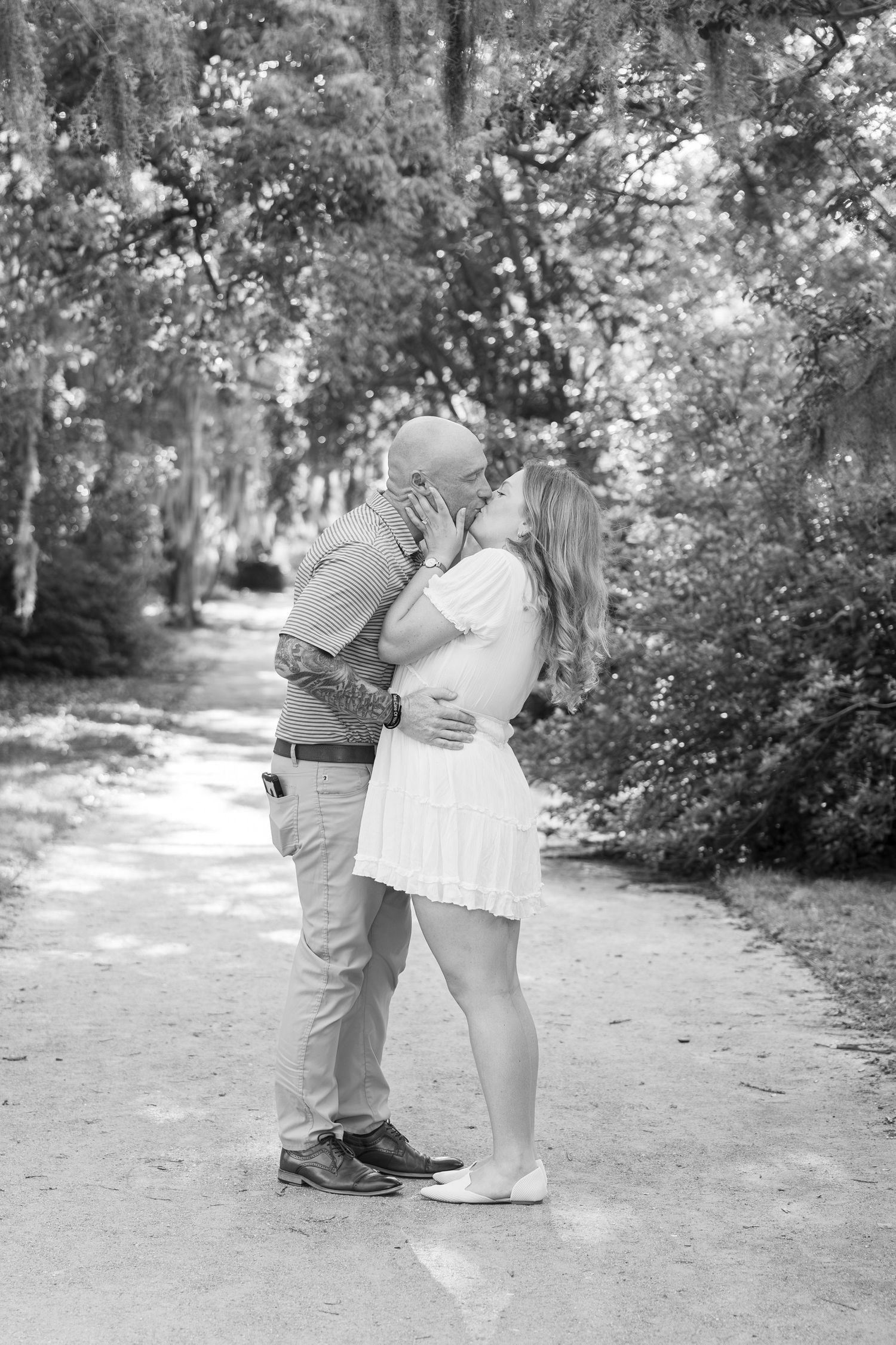 Black and white photo of a couple kissing on a tree-lined path, woman in white dress with visible baby bump.