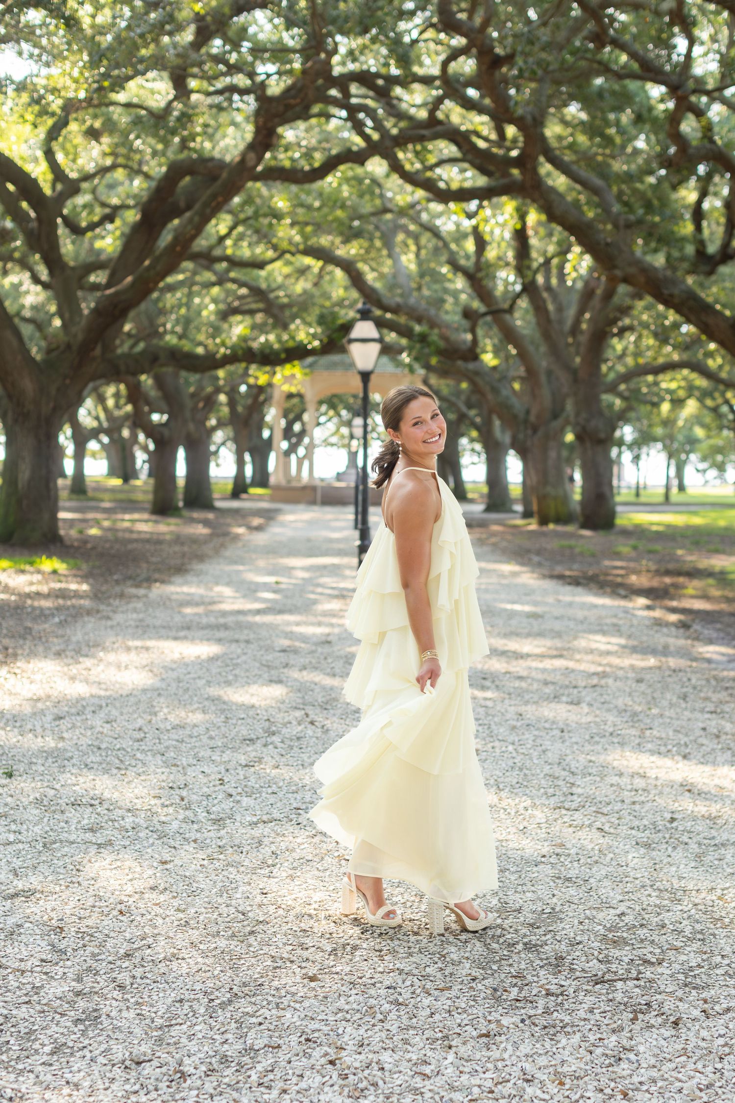 Bride in flowing white gown twirling on a tree-lined gravel path, smiling over her shoulder.