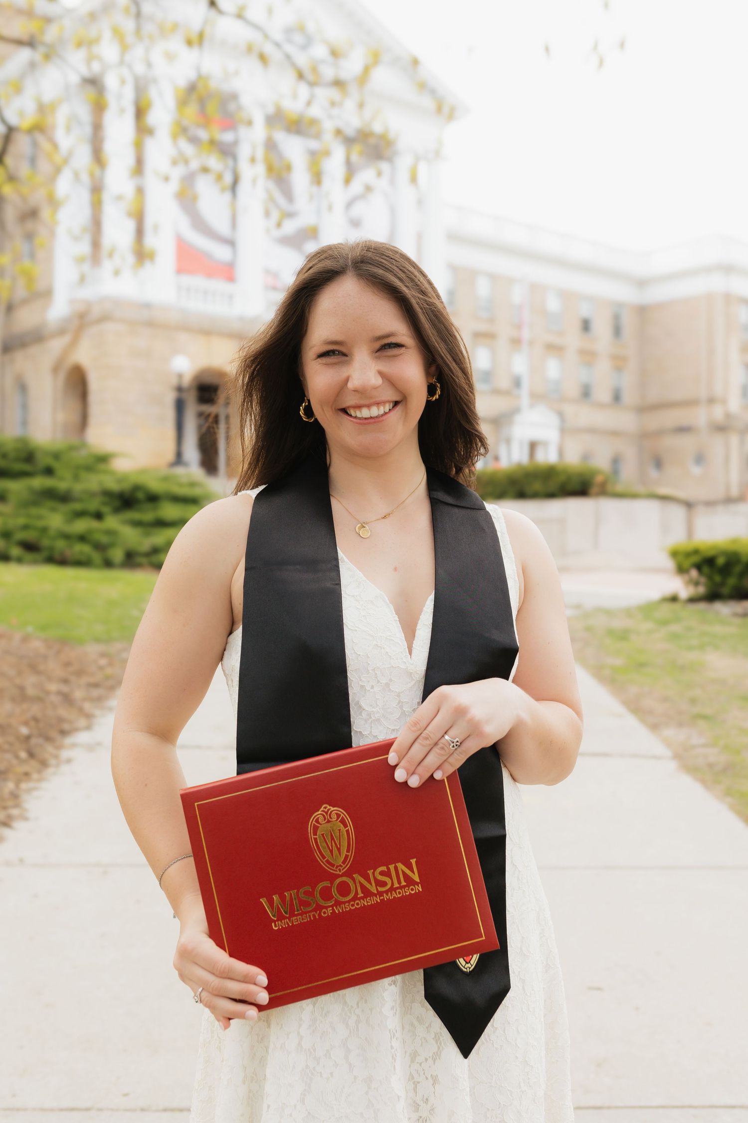 Graduate in white dress holds red Wisconsin diploma folder on campus grounds.