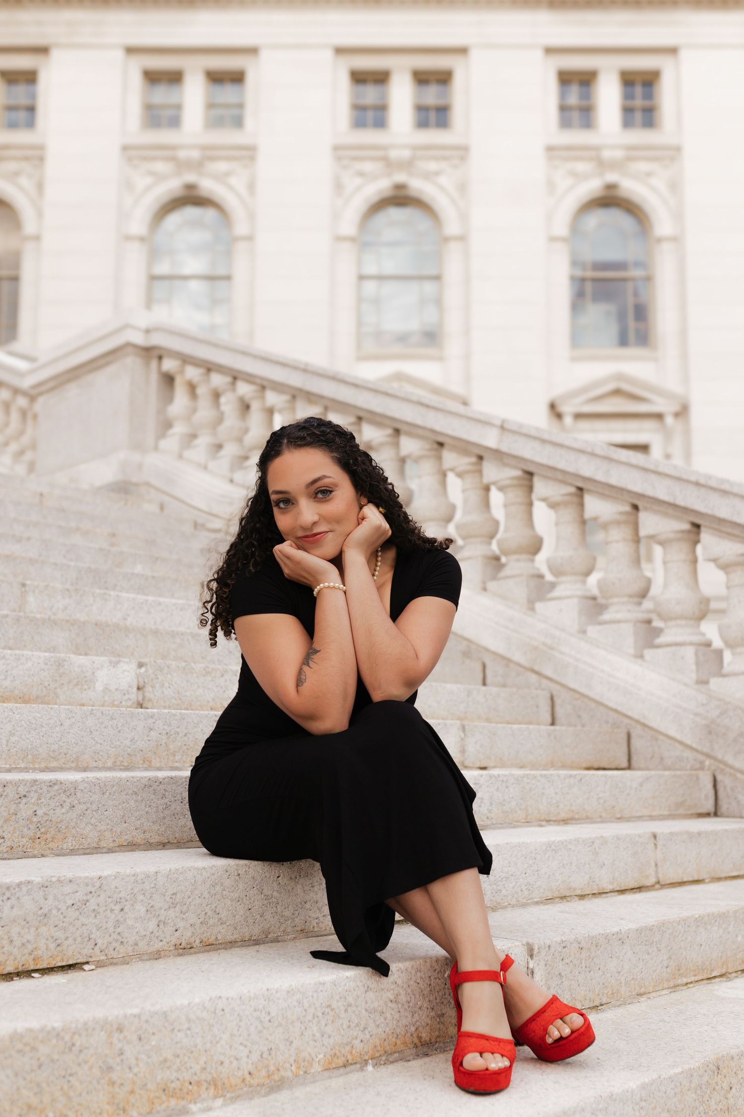 Graduate in a black dress and red heels poses on elegant stone steps of a historic building.