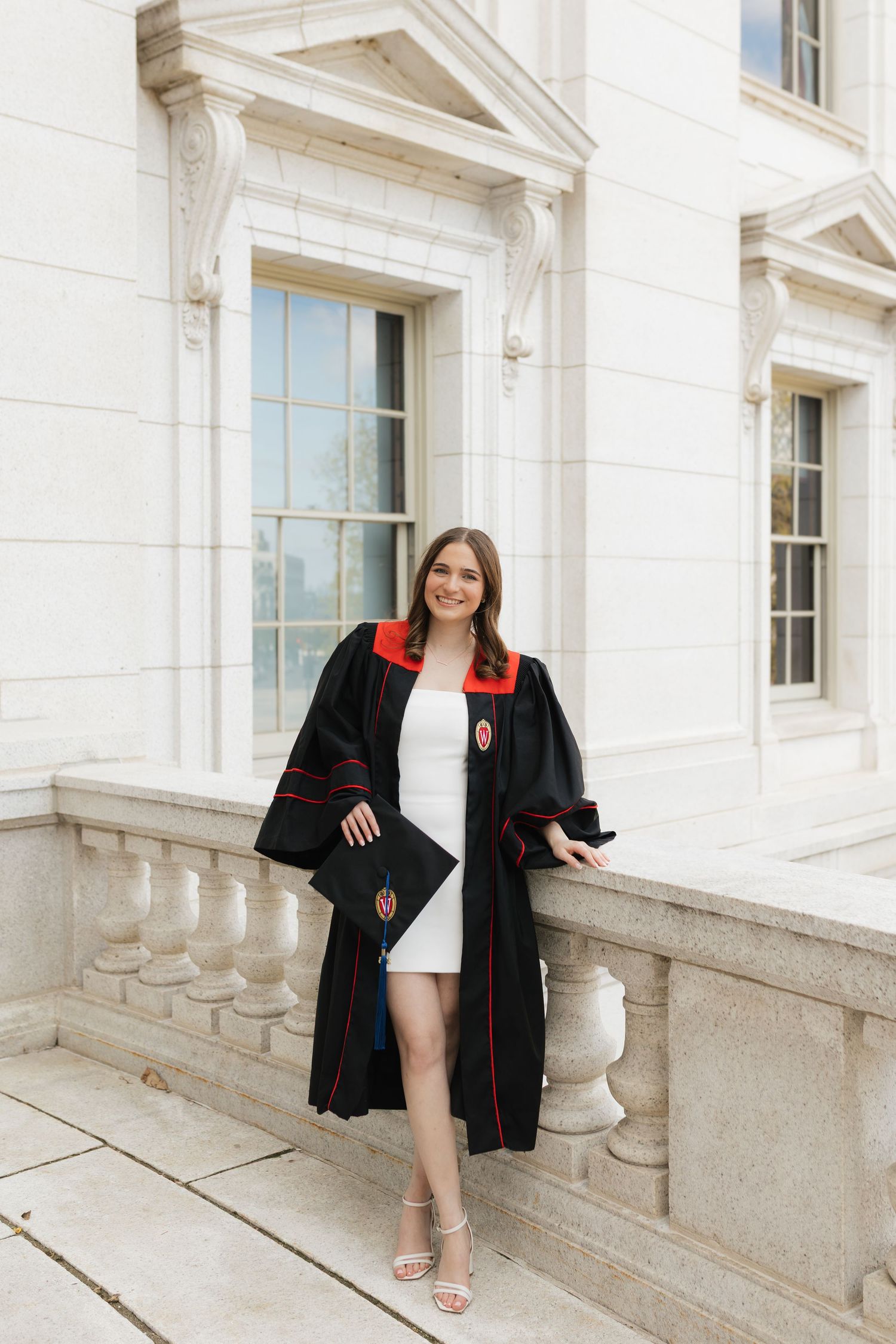 Graduate in black and red graduation gown poses on stone balcony of historic building.