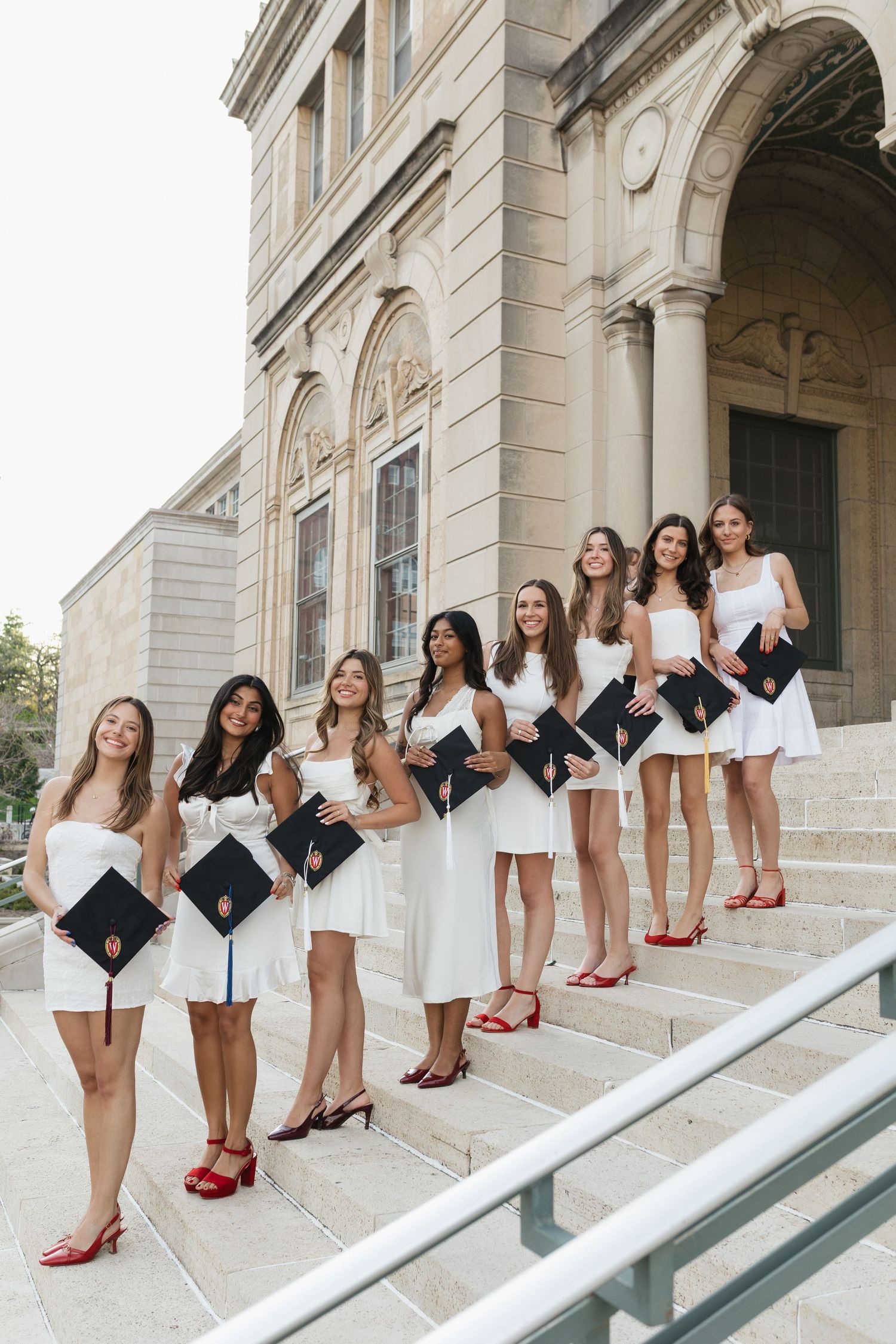 Graduates in white dresses and red heels pose on stone steps of university building.