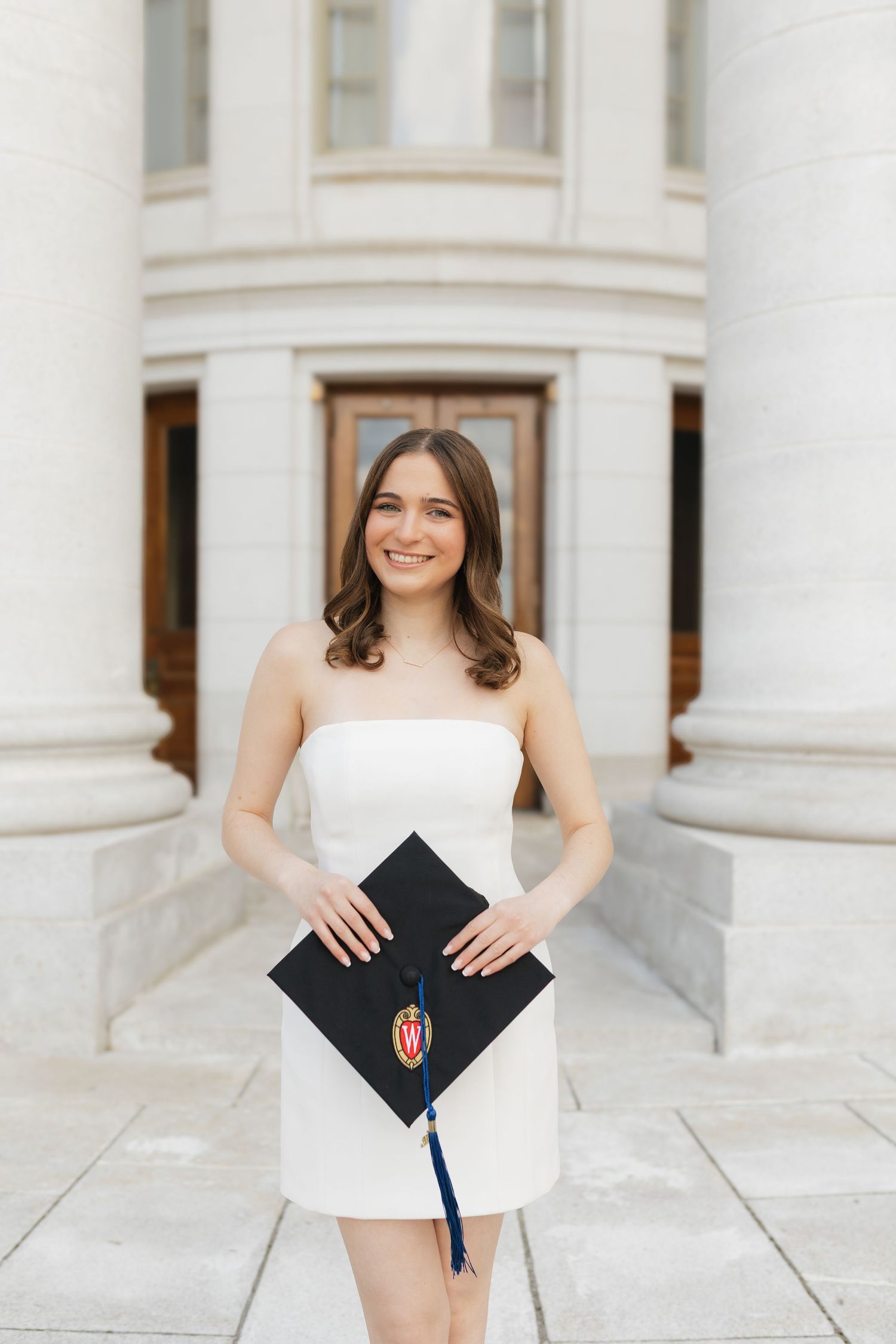 Graduate in white dress poses with diploma in front of classical white columns.